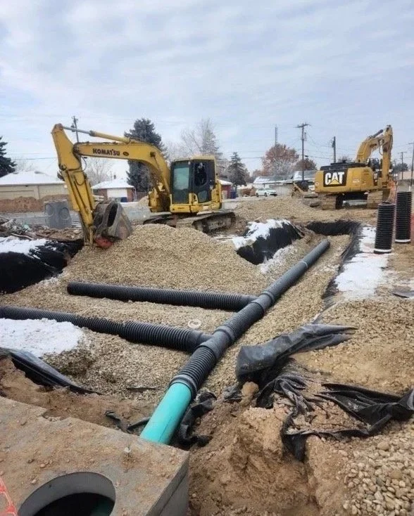 Construction site with two excavators, one labeled 'Komatsu' and the other 'Caterpillar,' working on an underground pipe installation in a snow-covered area.