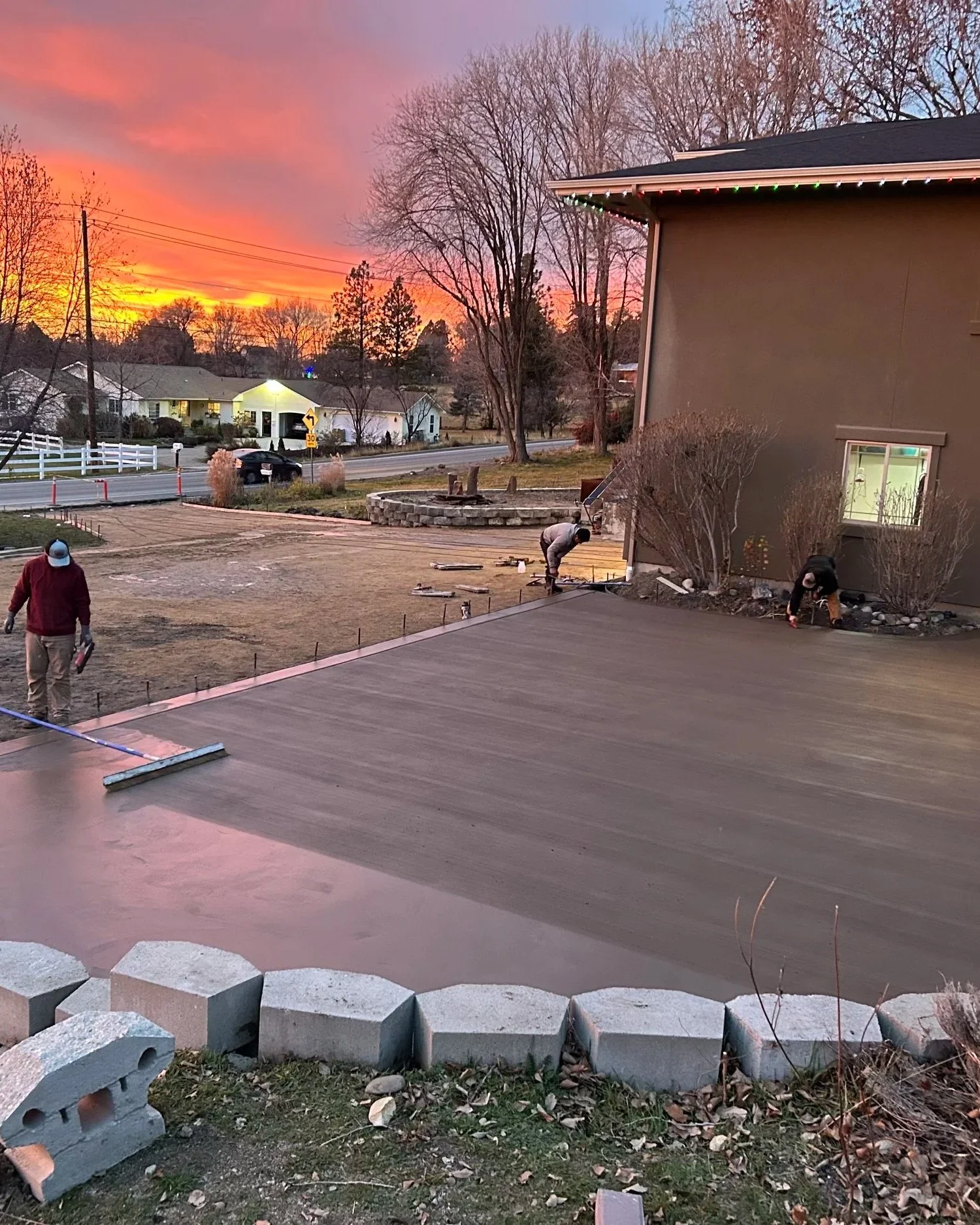 Construction workers working on a new concrete driveway or sidewalk at dusk or dawn, with a sunset or sunrise in the background, in a residential neighborhood.