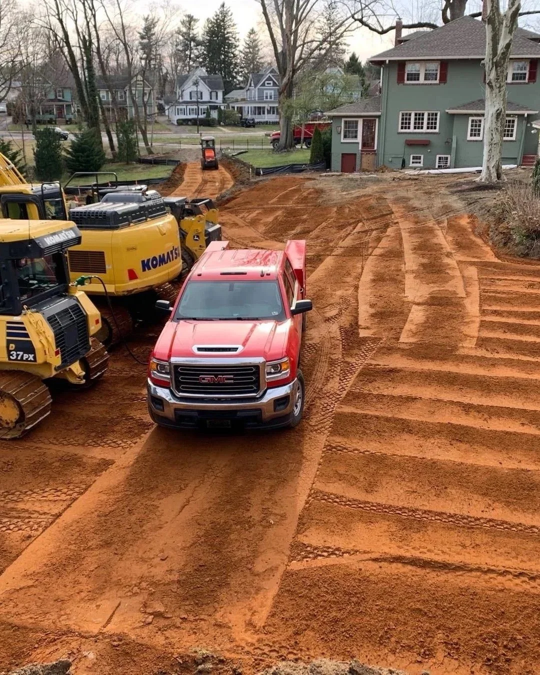 A construction site with a red GMC truck, yellow bulldozer, and other construction equipment on a dirt lot in a residential area. The area appears to be under development or grading.