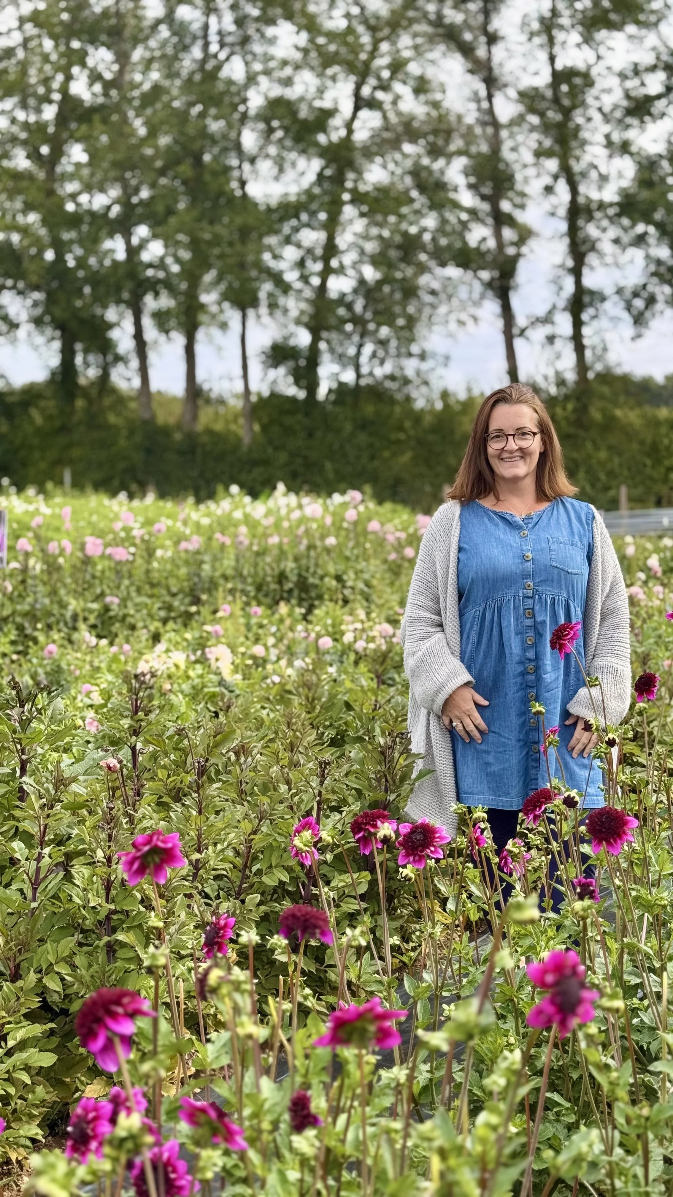 A woman smiling outdoors in a garden of pink and purple flowers, with trees and a cloudy sky in the background.