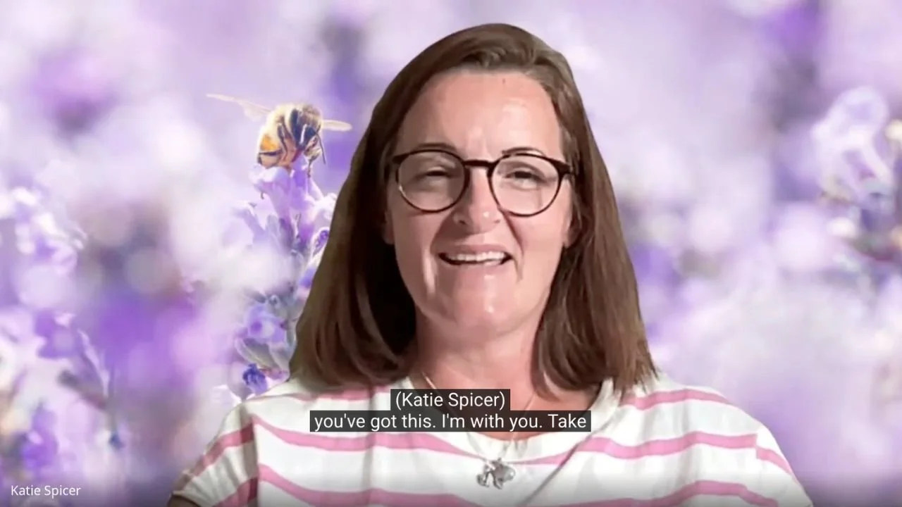 A woman, Katie Spicer, with shoulder-length brown hair and glasses, smiling in front of a purple floral background, wearing a pink and white striped shirt.