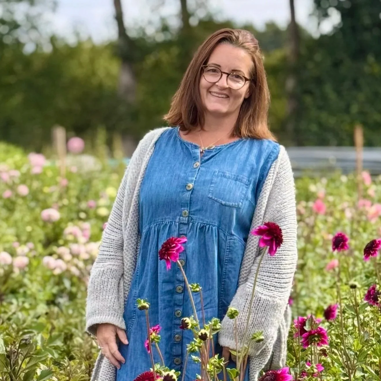 A woman with shoulder-length brown hair and glasses standing in a flower garden with pink and purple flowers.