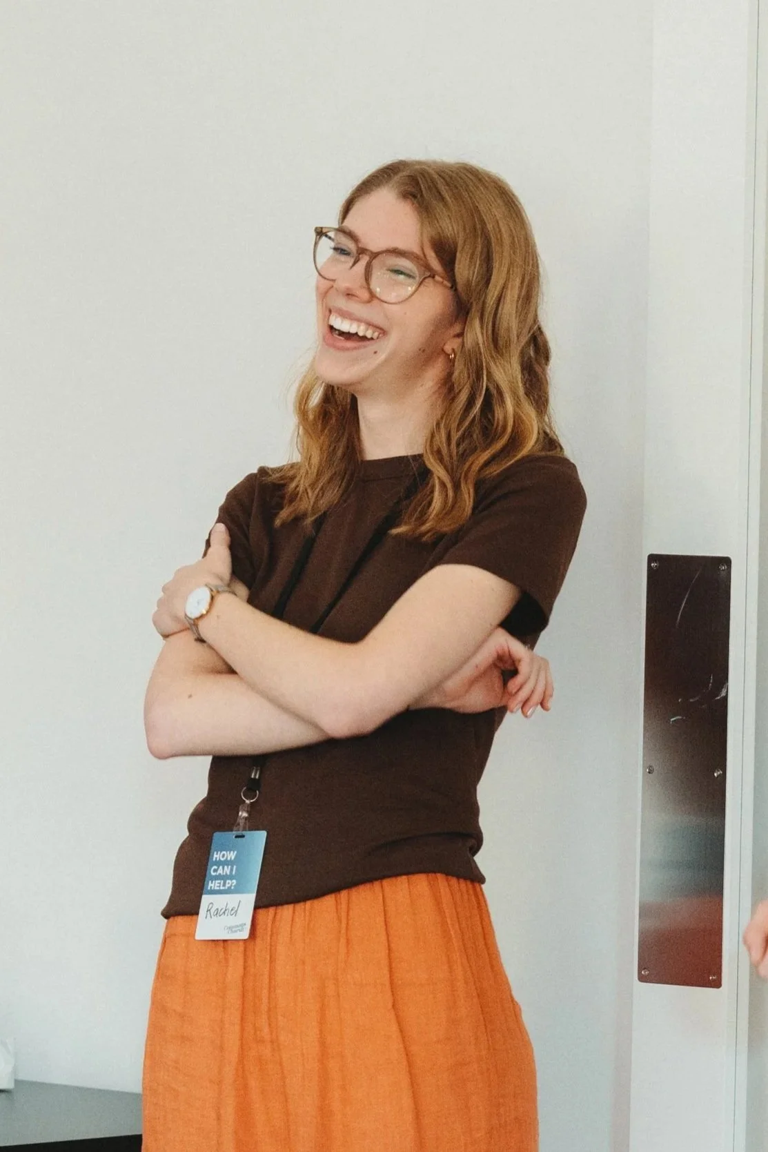 A woman with red hair and glasses, smiling and crossing her arms, wearing a brown shirt and orange skirt, with a name tag that says Rachel, in an indoor setting.