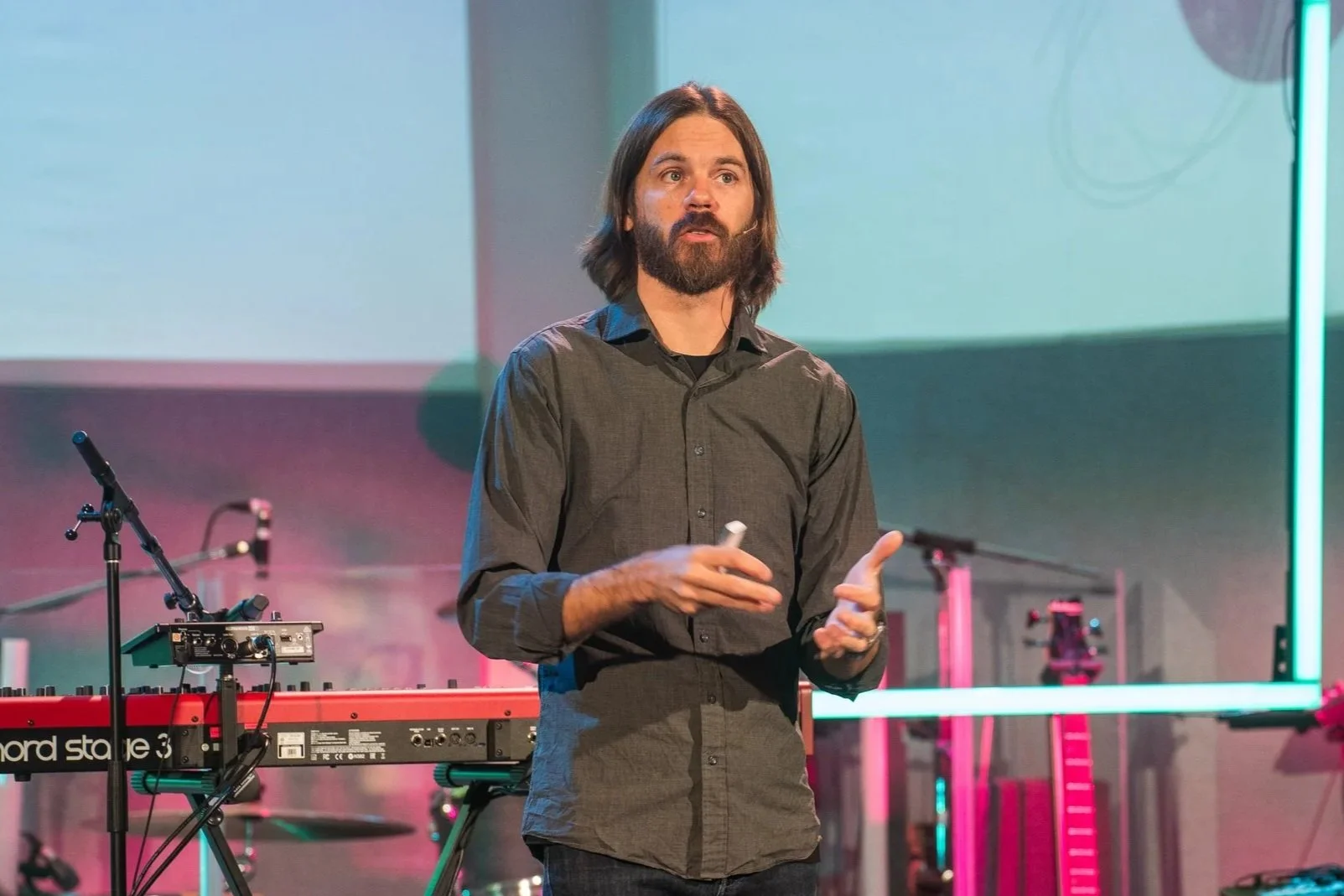 Man with shoulder-length dark hair and beard giving a presentation on stage with electronic musical equipment behind him, including a red keyboard and sound mixer, and colorful stage lighting.