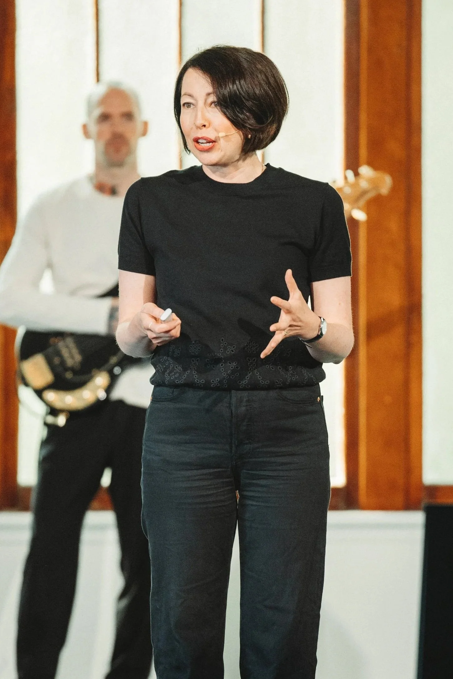 A woman with short dark hair speaking in front of a microphone, gesturing with her hand, in a setting with wooden paneling and a man playing guitar in the background.