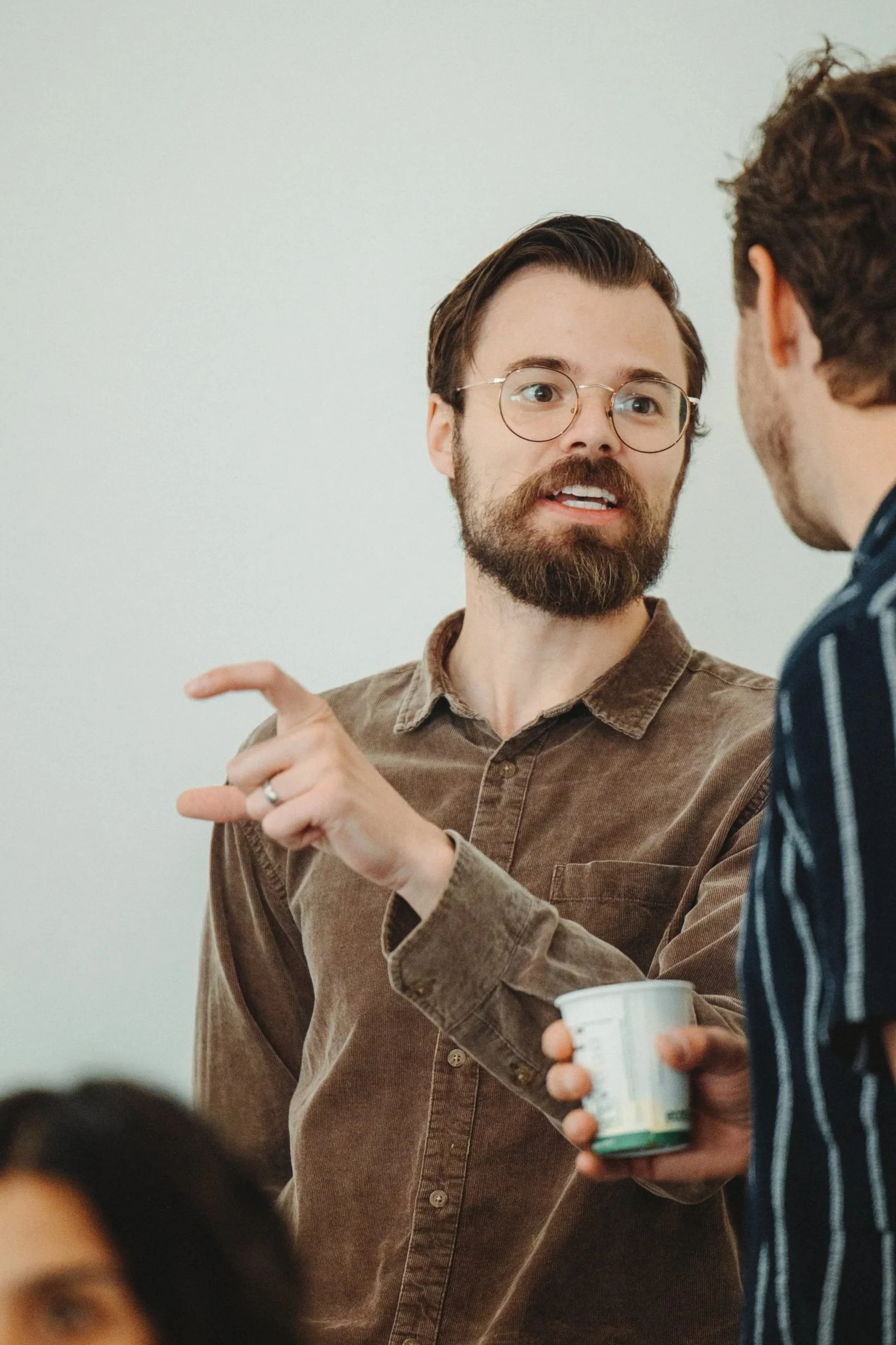 Two men engaged in conversation, one with glasses and a beard holding a paper cup, the other partially visible from behind, wearing a striped shirt.