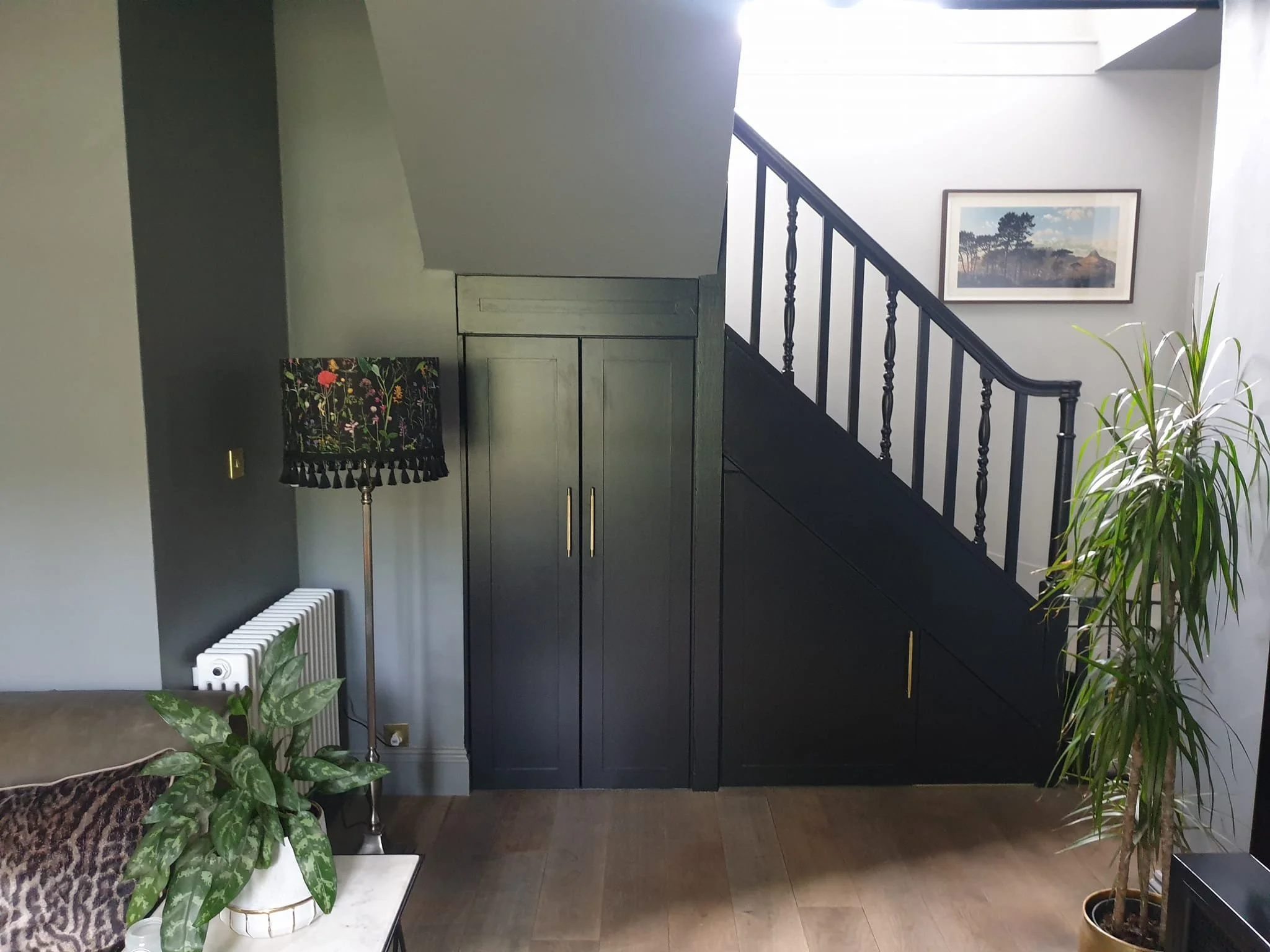 Interior view of a modern house with a staircase, framed landscape picture on the wall, various potted plants, a colorful floral lampshade, and a radiator.