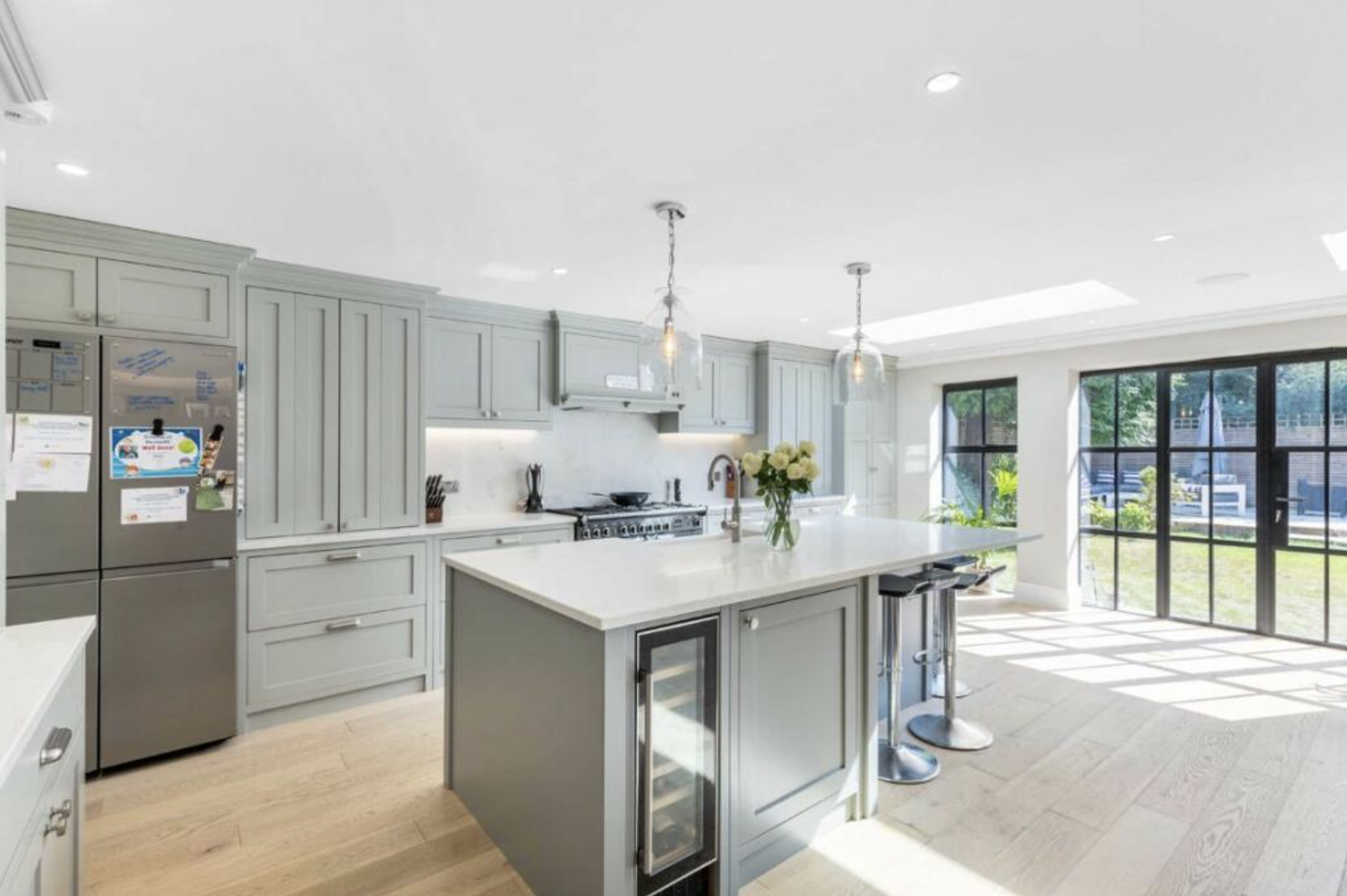 Bright modern kitchen with light gray cabinets, a white island, and large windows showing an outdoor area with trees.