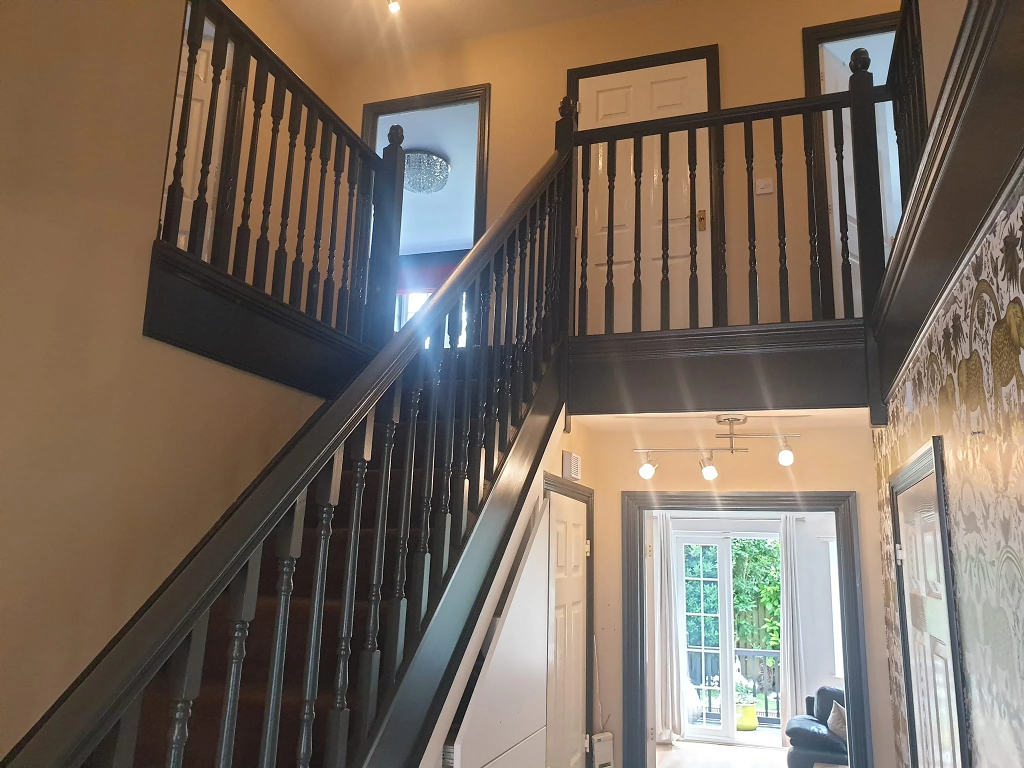 View of an interior staircase with dark wooden balustrades, leading to a small upper landing with a white door, in a house with beige walls, a patterned wallpaper border, and a large window at the bottom of the stairs revealing trees outside.