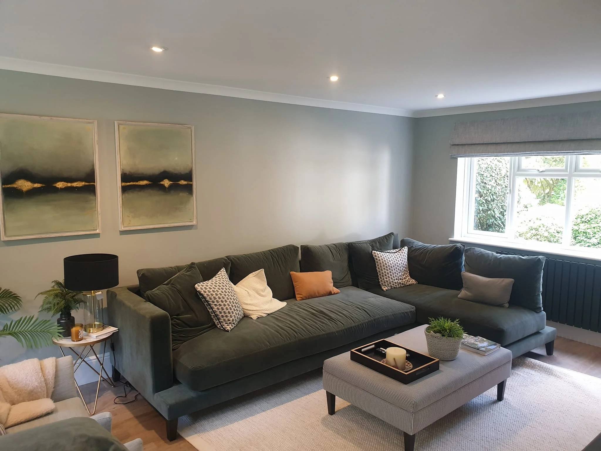 Living room with a large green sofa decorated with various patterned throw pillows, a coffee table with a plant and a tray, a side table with a black lamp, and a window with shades.
