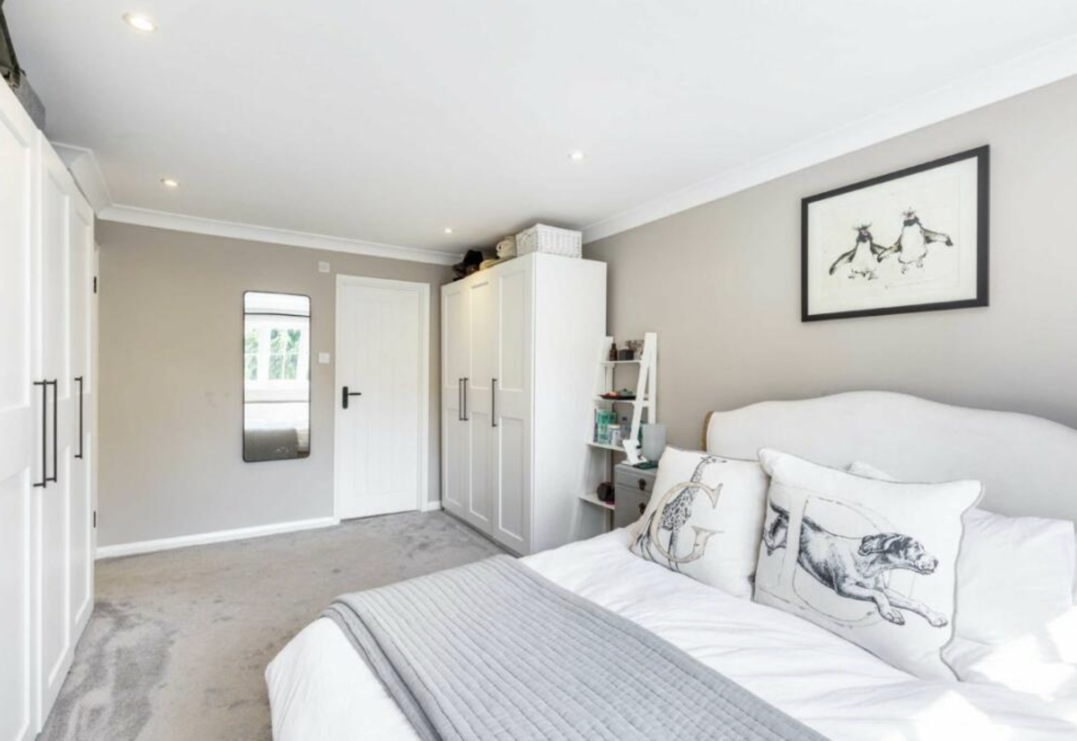 A cozy bedroom with a white bed featuring black and white animal print pillows, a framed art piece of two deer on the wall, white wardrobes, and a small white shelving unit. There is a window and a door in the room.