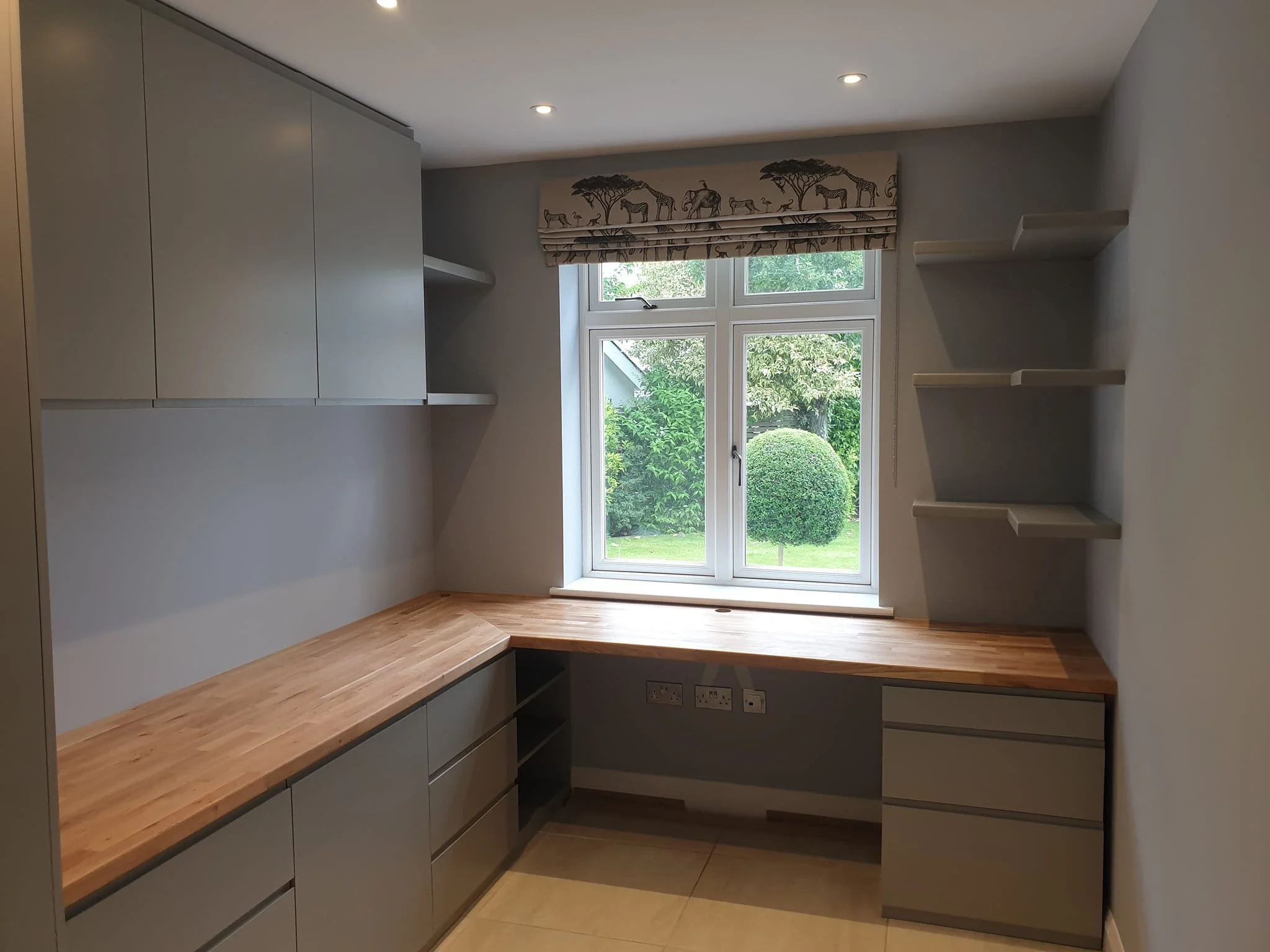 Modern kitchen with wooden countertops, white cabinets, large window with safari animal print valance, and green trees visible outside.