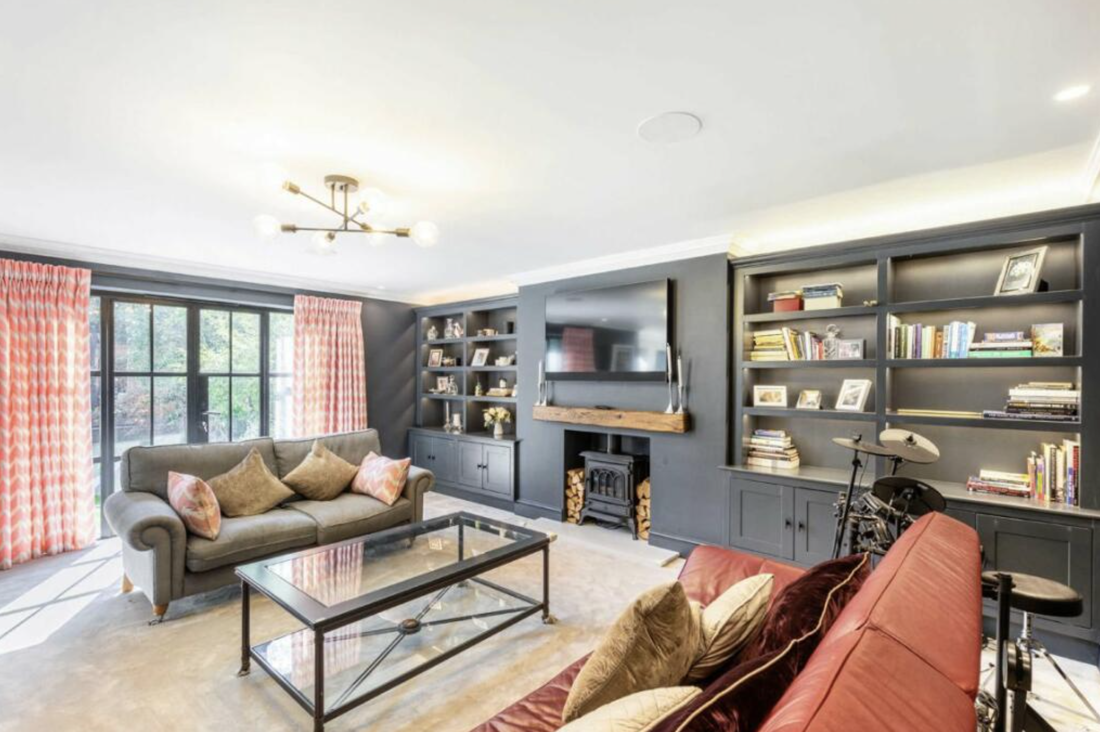 Living room with gray sofa, red and patterned pillows, glass coffee table, large window with pink curtains, built-in bookshelves, flat-screen TV, and a chandelier.