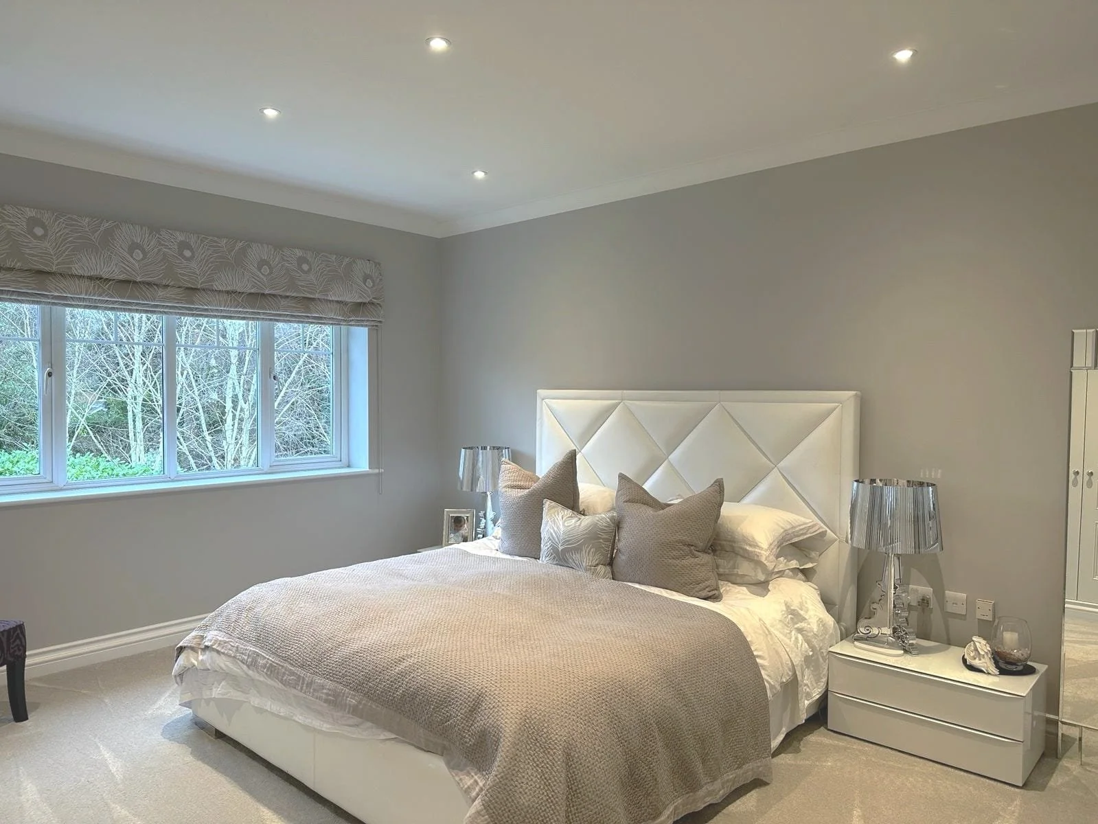 A modern bedroom with a large bed, multiple pillows, a white headboard, bedside tables with lamps, and a window with patterned curtains, overlooking trees outside.