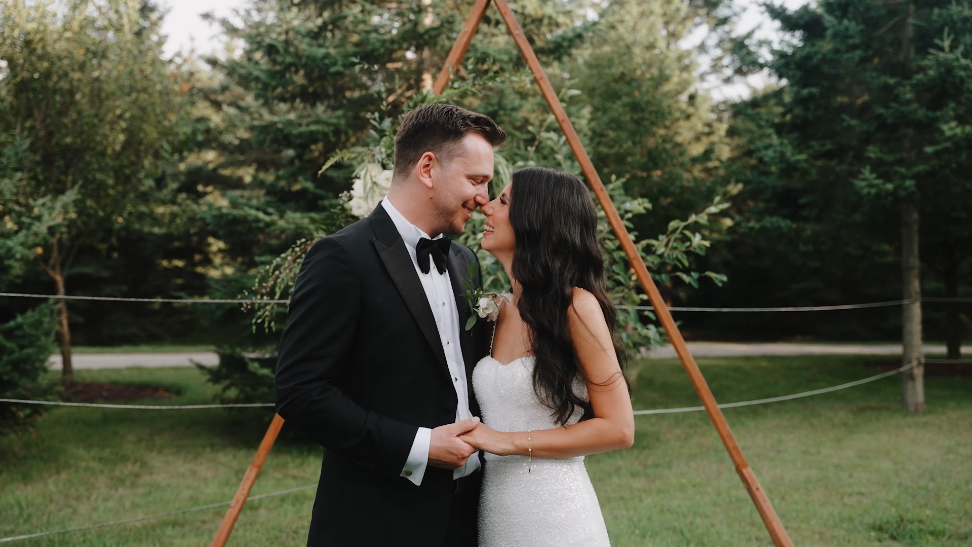 A newlywed couple in wedding attire sharing an intimate moment outdoors, with trees and a wooden triangular wedding arch decorated with white flowers and greenery in the background.