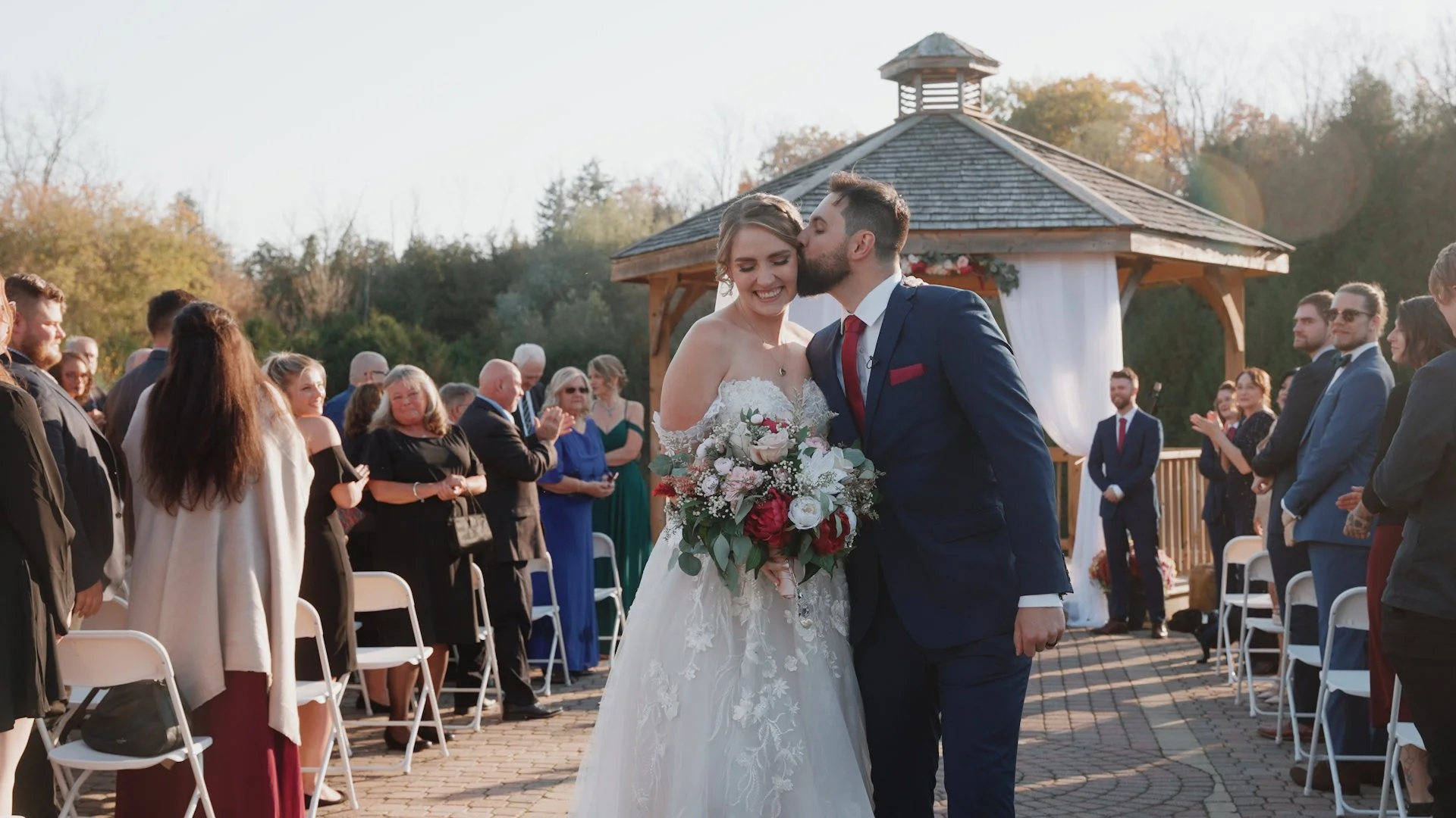 A bride and groom on their wedding day, with the groom kissing the bride's forehead, surrounded by wedding guests outdoors during daytime.