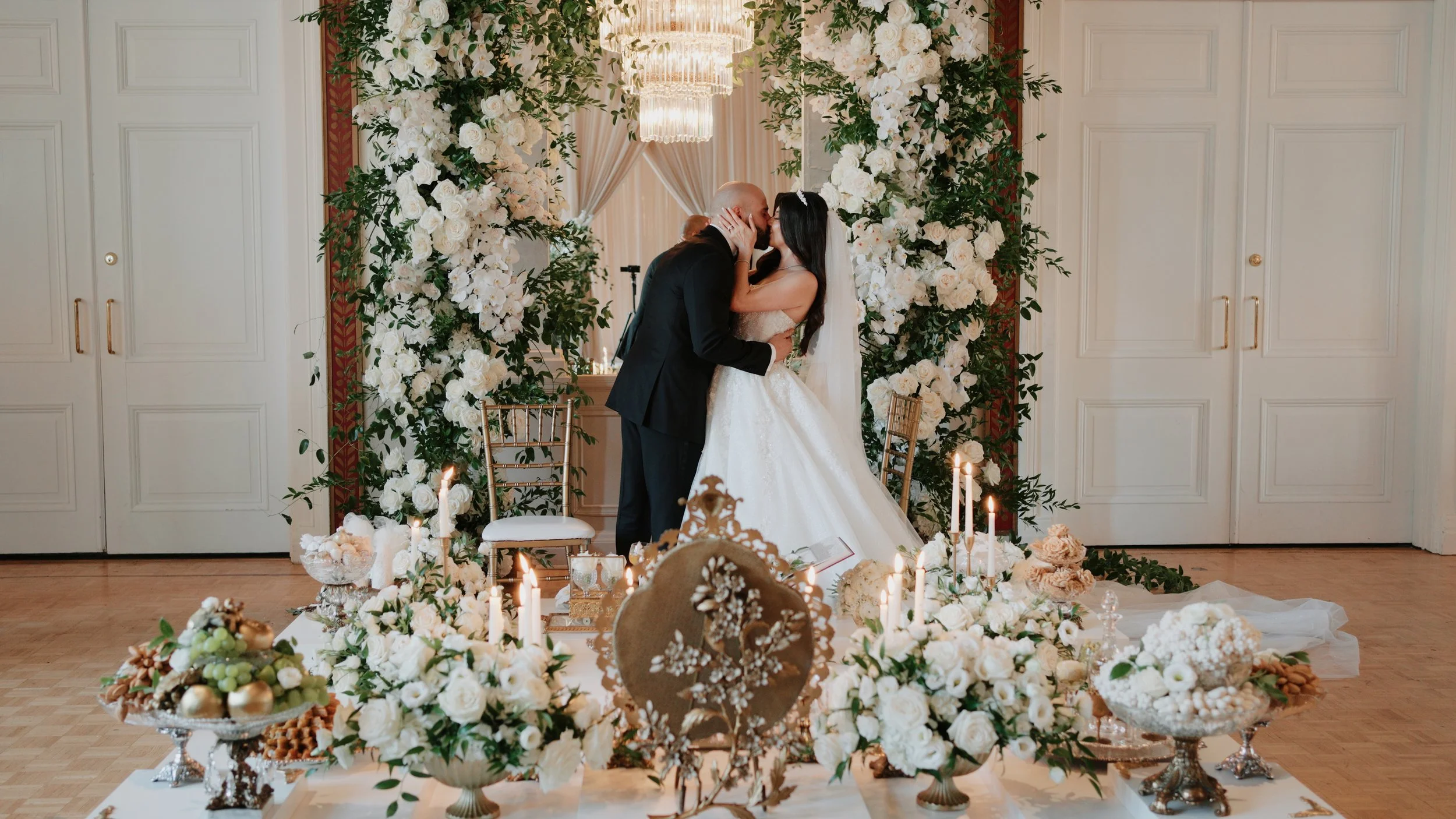A bride and groom kissing at their wedding ceremony, surrounded by an elaborate floral arch and decorated with candles and flower arrangements.