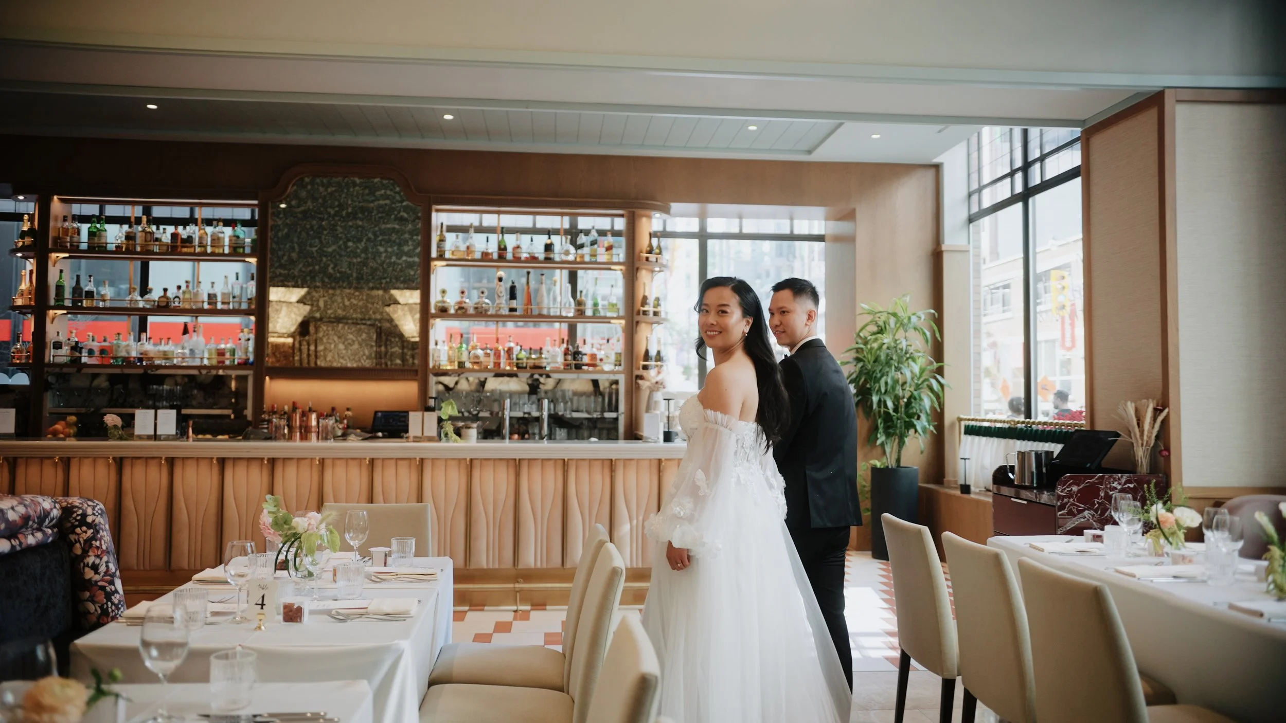 Bride and groom standing in a restaurant or banquet hall decorated for a wedding, with elegantly set tables and a bar in the background.