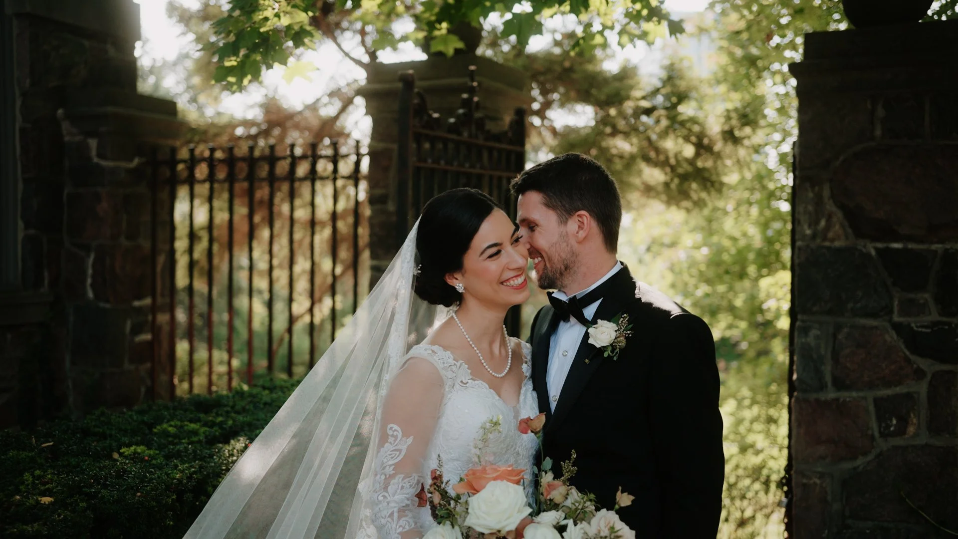 Bride and groom smiling close together outside, with trees and a black wrought iron gate in the background.