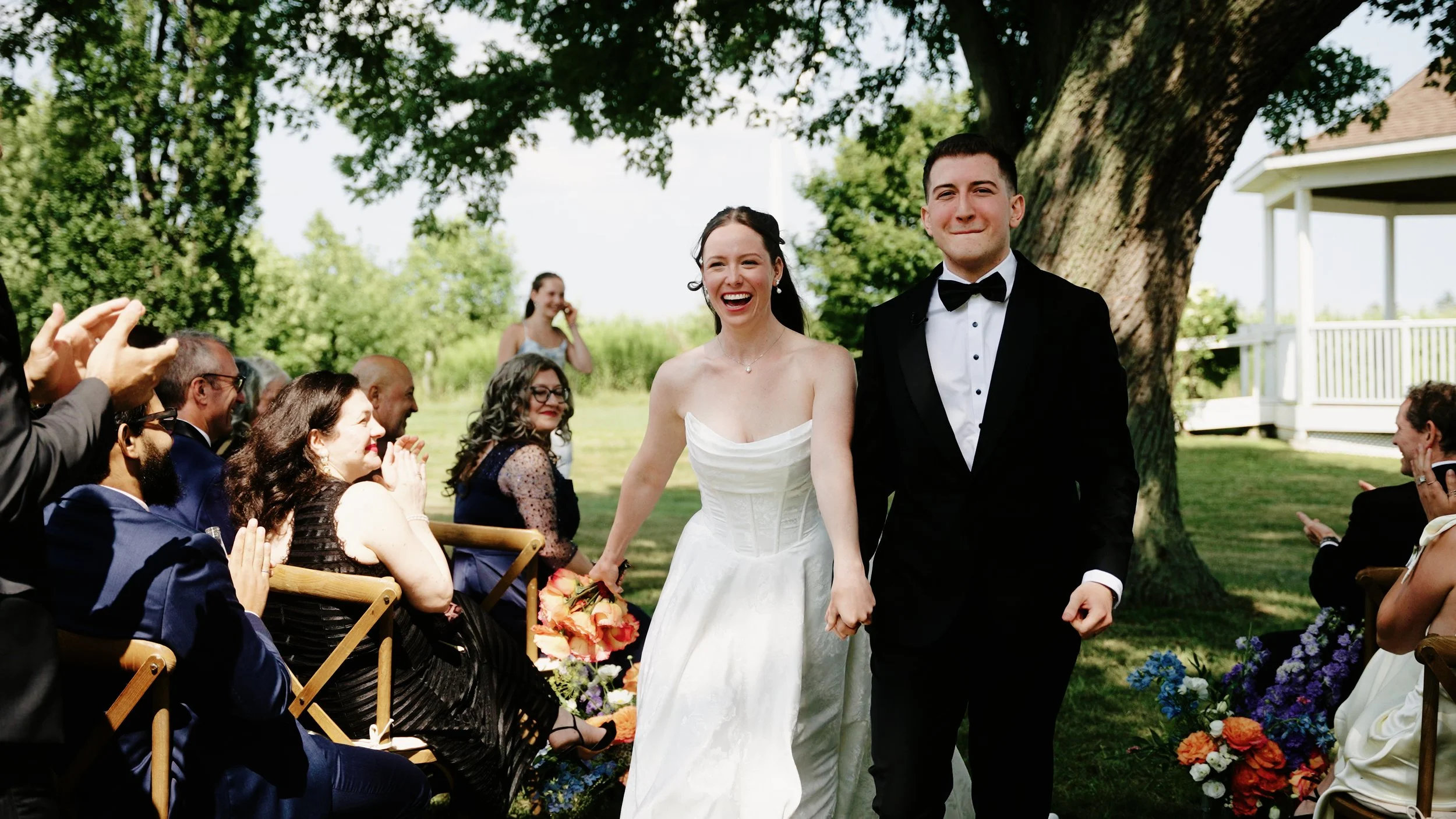 A bride and groom walk hand in hand down the aisle outdoors, smiling, with wedding guests seated on either side, some clapping, atop a grassy area under a large tree.