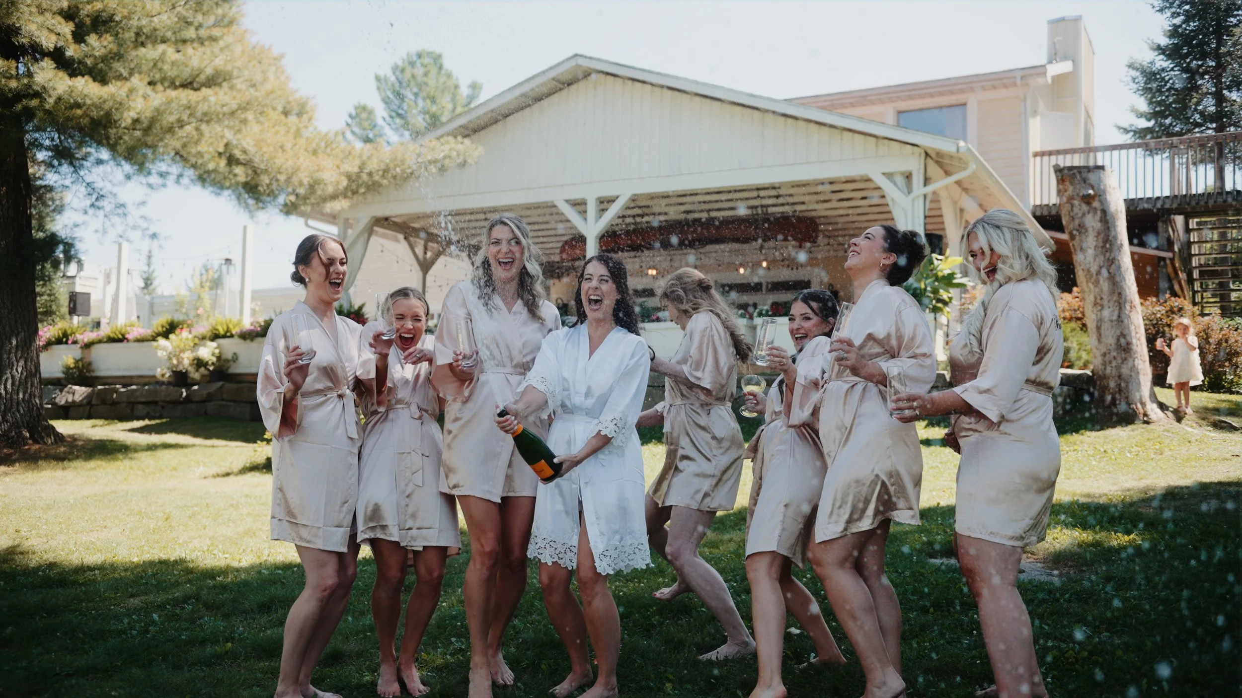 Group of women in matching satin robes celebrating outdoors with drinks and champagne, laughing and having fun in a garden with trees and a house in the background.