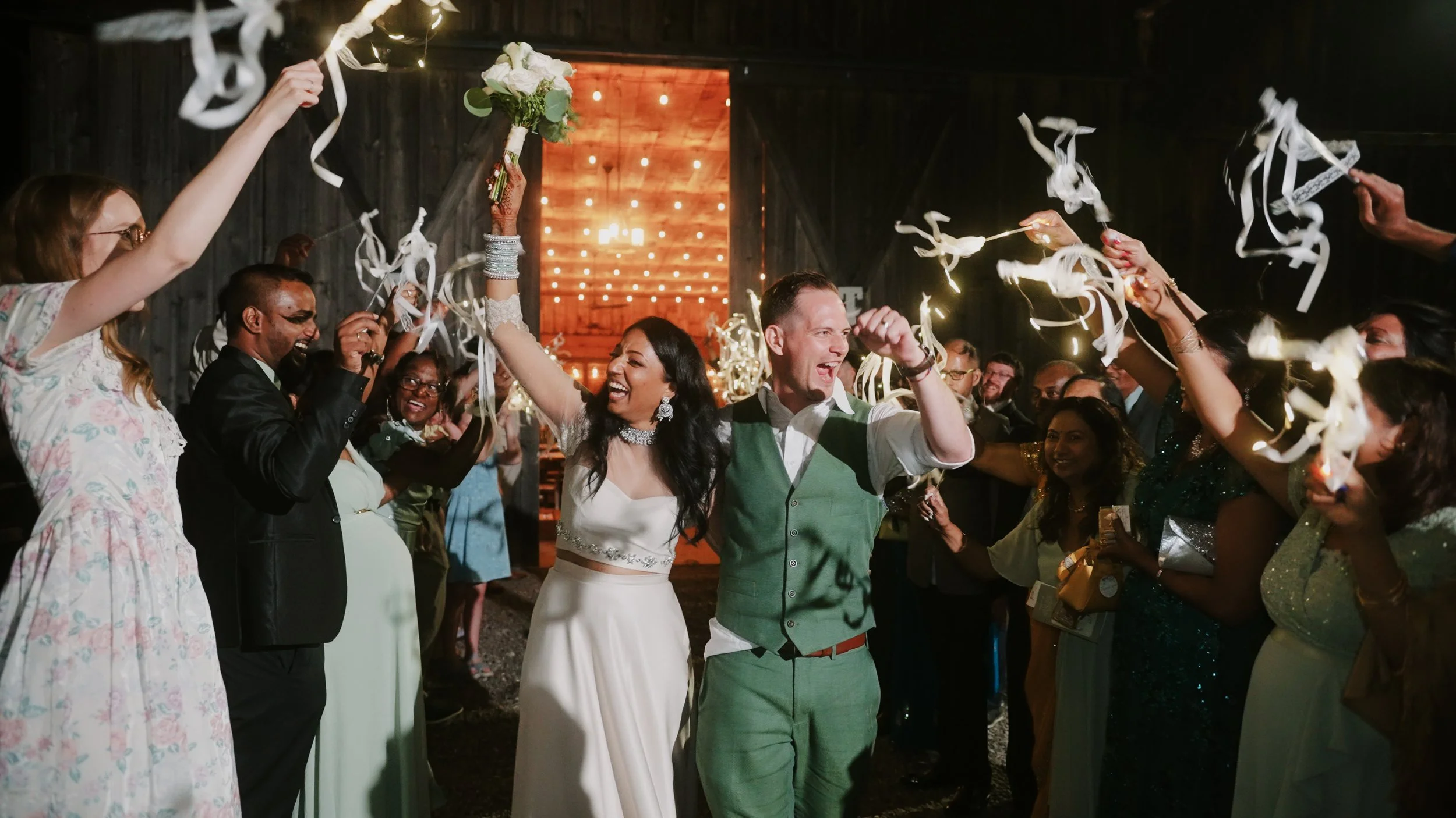 Wedding celebration with the bride and groom celebrating among guests, holding sparklers, in a rustic barn setting.