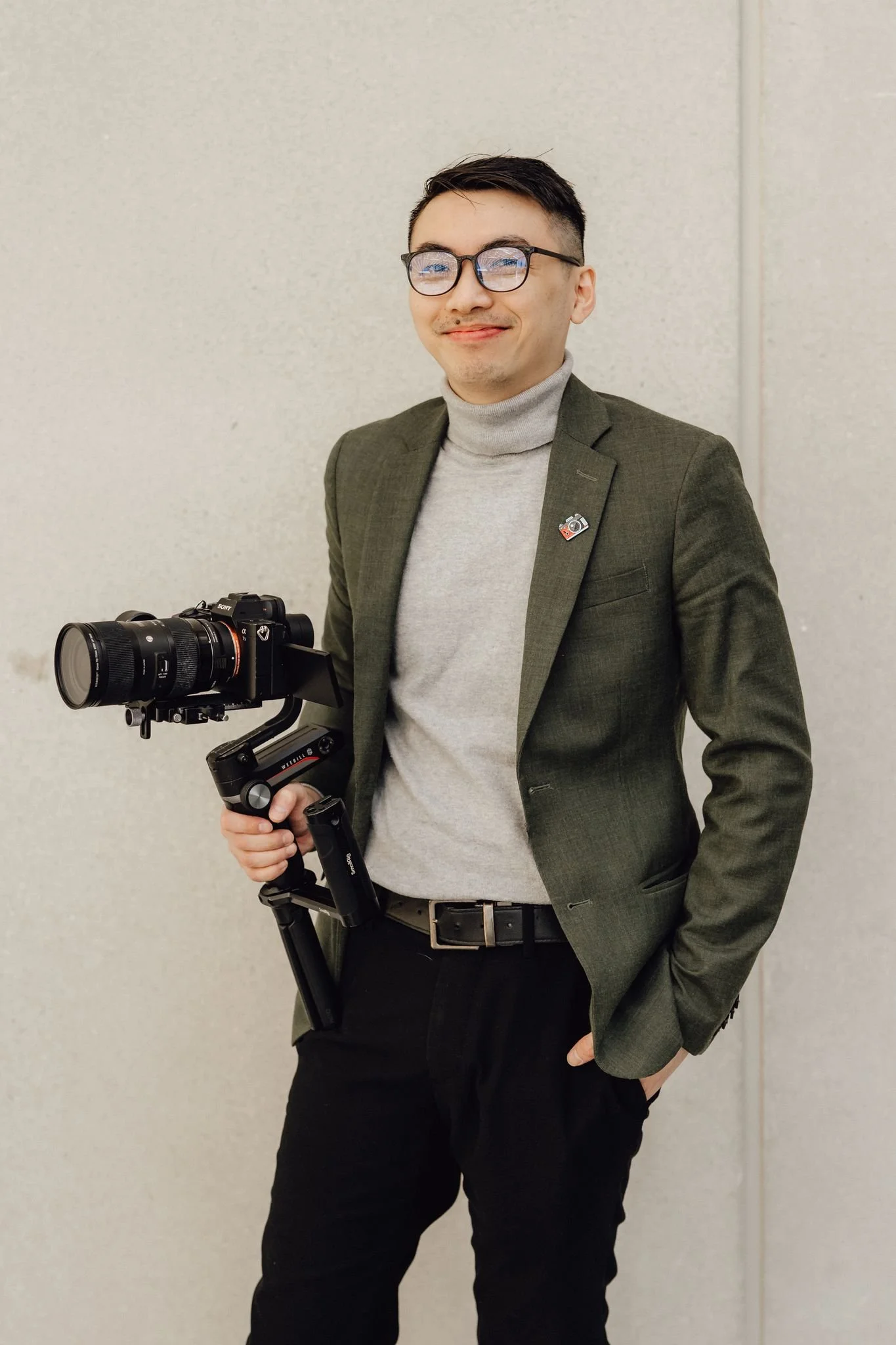 A young man with glasses, a slight smile, and dark hair, dressed in a gray turtleneck and a green blazer, holding a professional camera with a stabilizer against a light plain wall.
