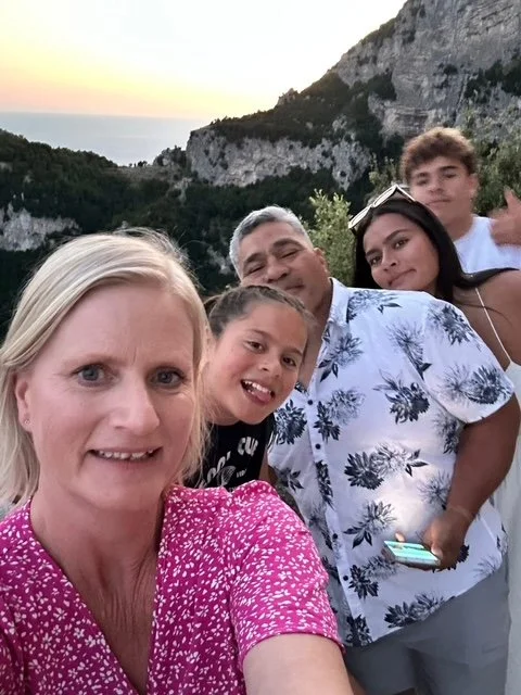 Group of five people taking a selfie outdoors at sunset, with a rocky hillside and trees in the background.