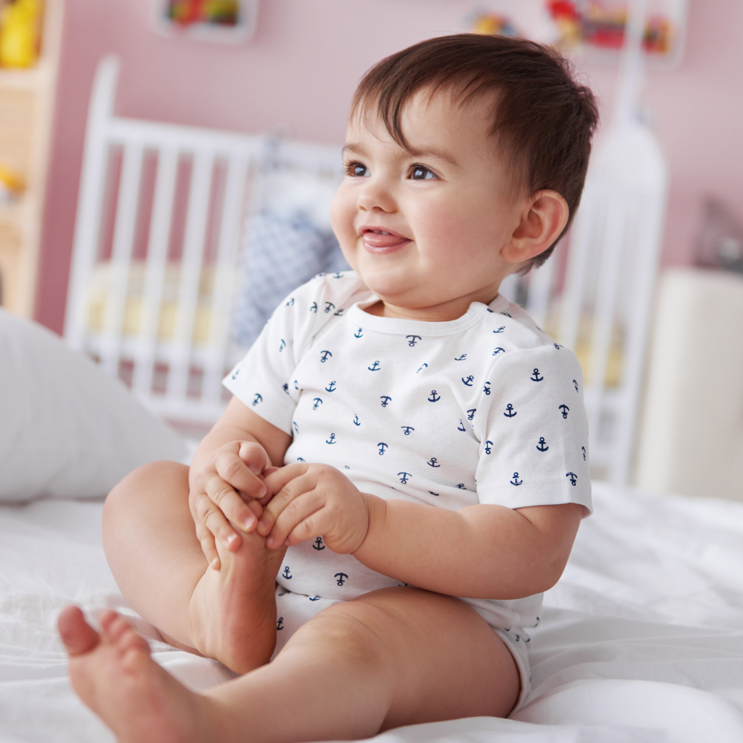 Baby sitting on a bedroom floor holding their right foot with cot in the background.  Child wearing white bodysuit with small blue anchors all over