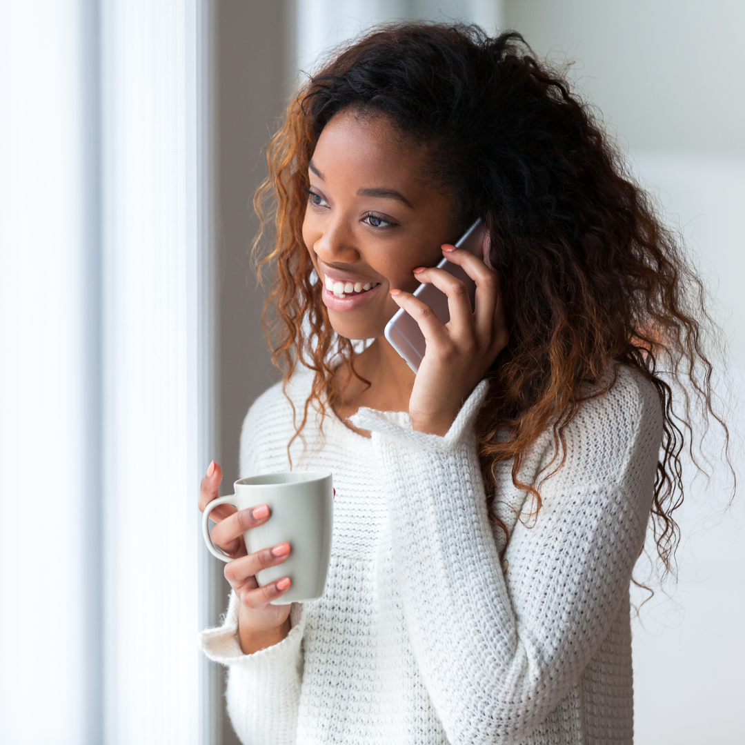 Smiling woman with curly hair talking on a phone while holding a white mug, near a bright window.