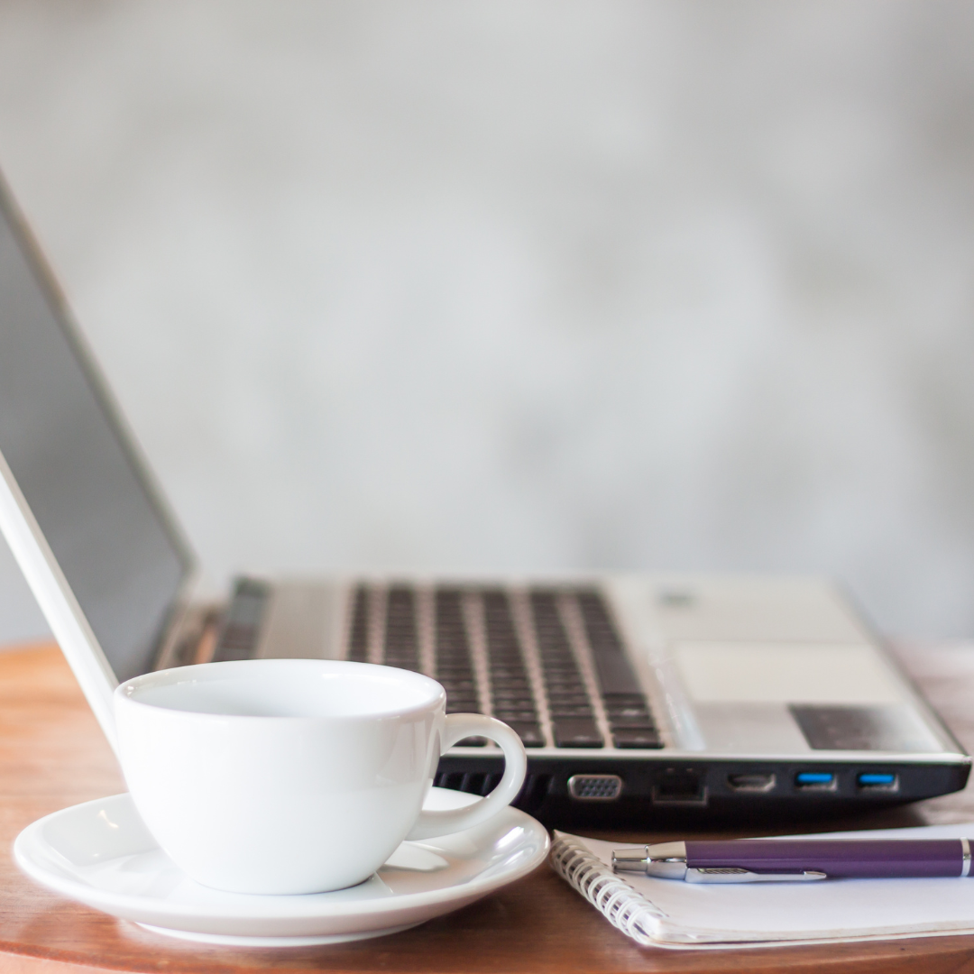 White coffee cup with saucer, spiral notepad, pen, and a silver laptop on a wooden surface.