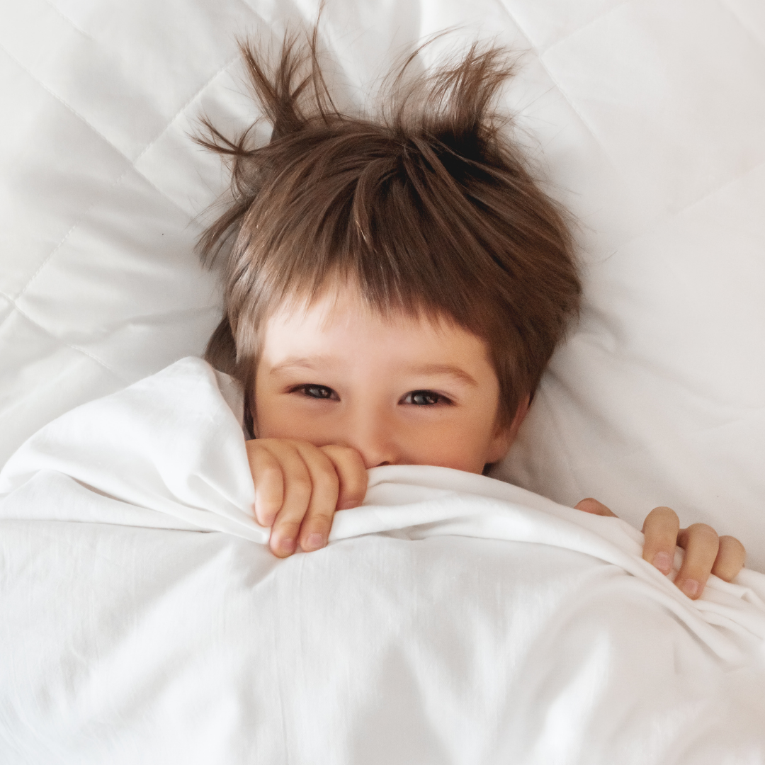 A young boy with tousled brown hair peeking out from under white bedsheets.