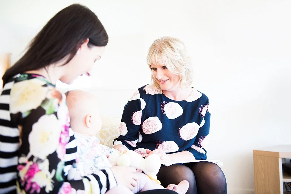 Two women and a baby sitting on a couch in a bright room, engaging in a cheerful conversation.
