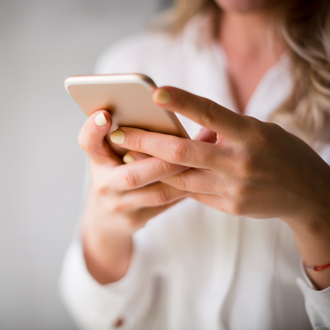 A woman with blonde hair, wearing a white blouse, is using a smartphone, holding it with both hands.