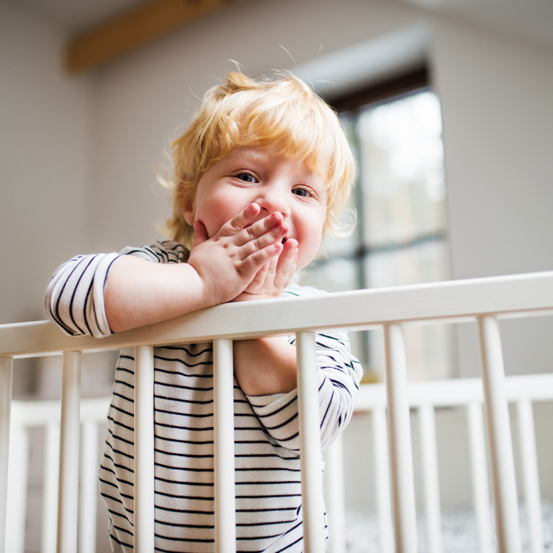 Toddler wearing white long sleeved top with blue stripes leaning over top of cot holding both hands to their mouth smiling in the background room has white walls with black window frame