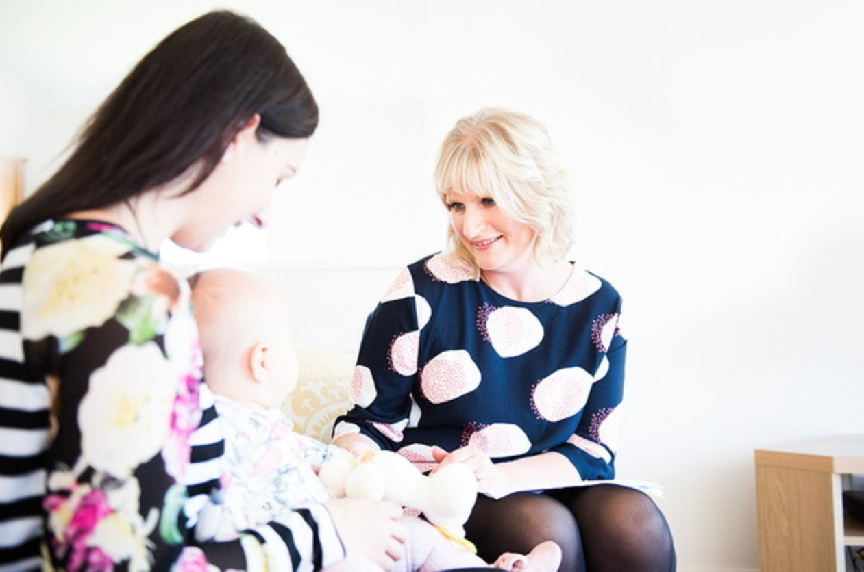 A woman with dark hair and a floral top holds a baby, while another woman with blonde hair and a patterned dress sits nearby, smiling at the baby in a bright, modern room.
