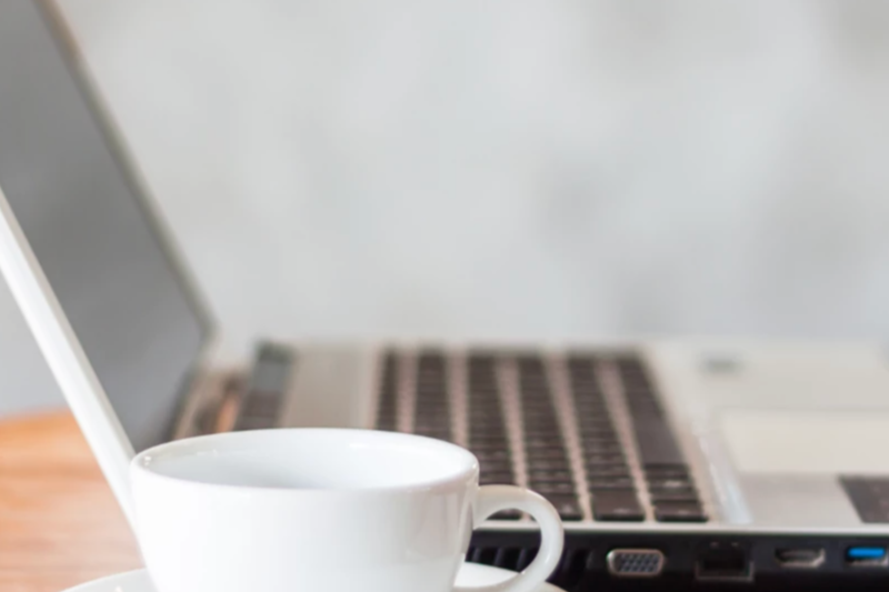 Close-up of a white coffee cup in front of a laptop on a wooden desk.