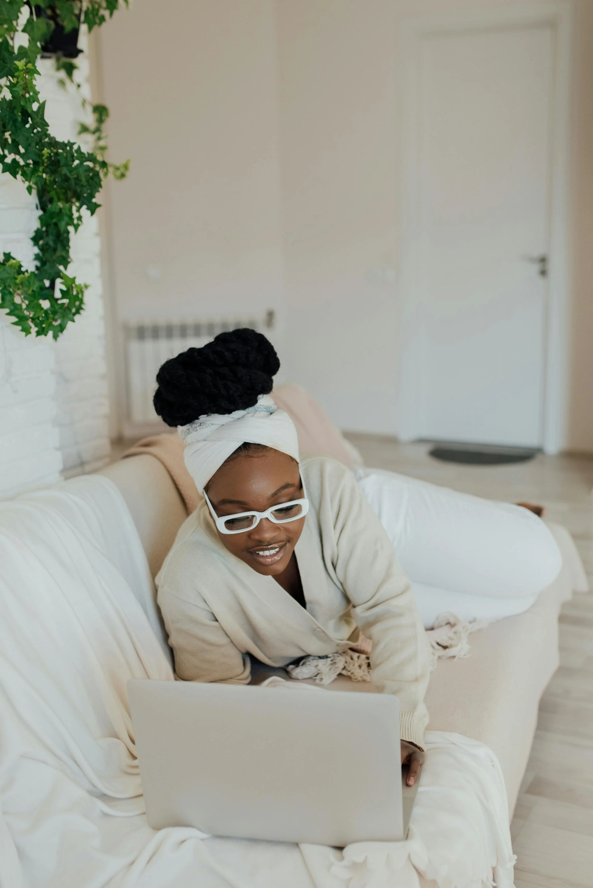 A black woman in a beige cardigan, white pants, white glasses, an afro and headwrap lounges on a couch with a computer. Online therapy for black women in Pasadena. Set boundaries, Find Peace