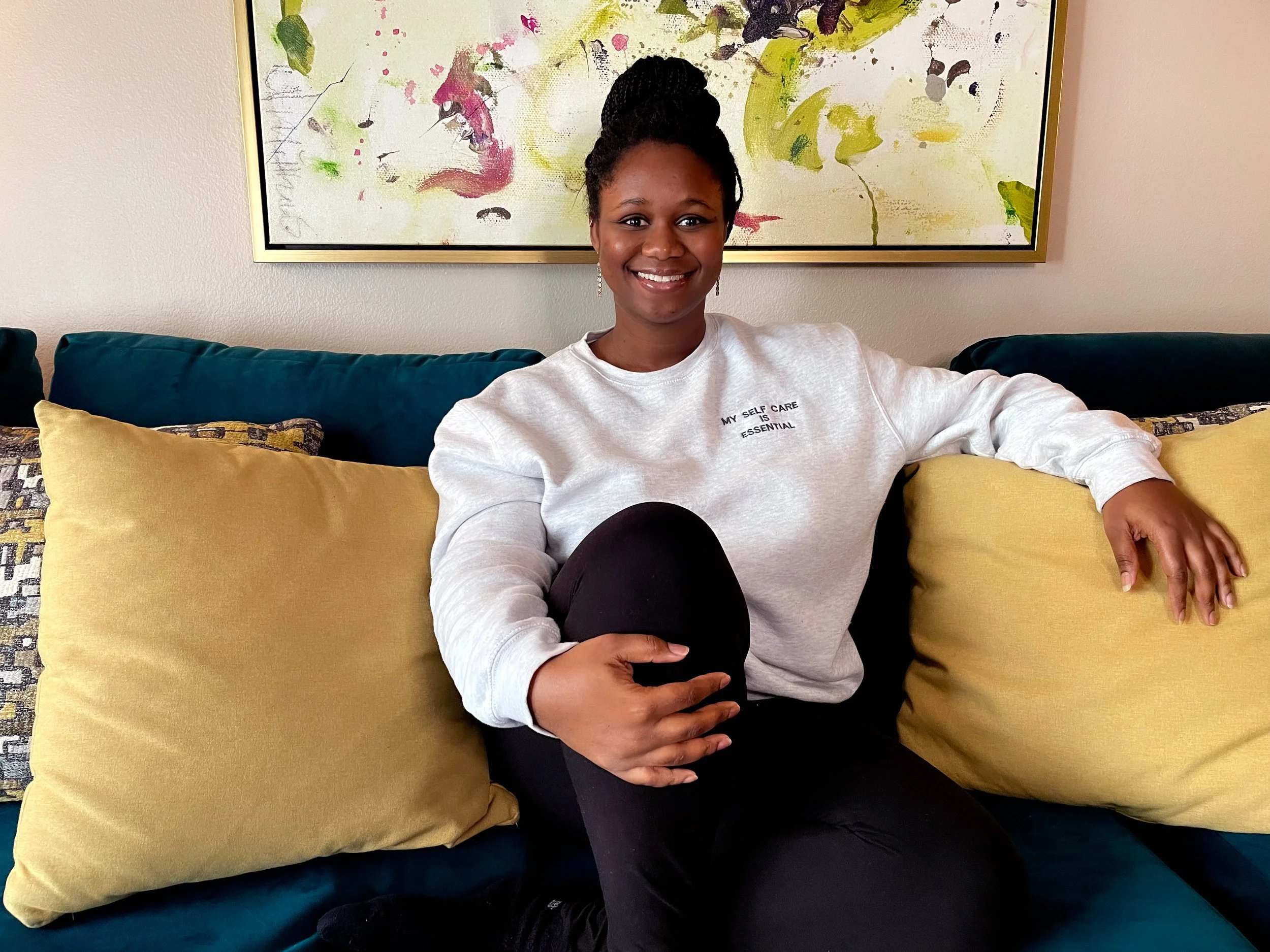 Crystal Clark, a Black in Therapist in Los Angeles sits on a green couch with yellow pillows. with an abstract painting in the background. She wears leggings and a grey sweatshirt that states "my self care is essential. Therapy for better boundaries