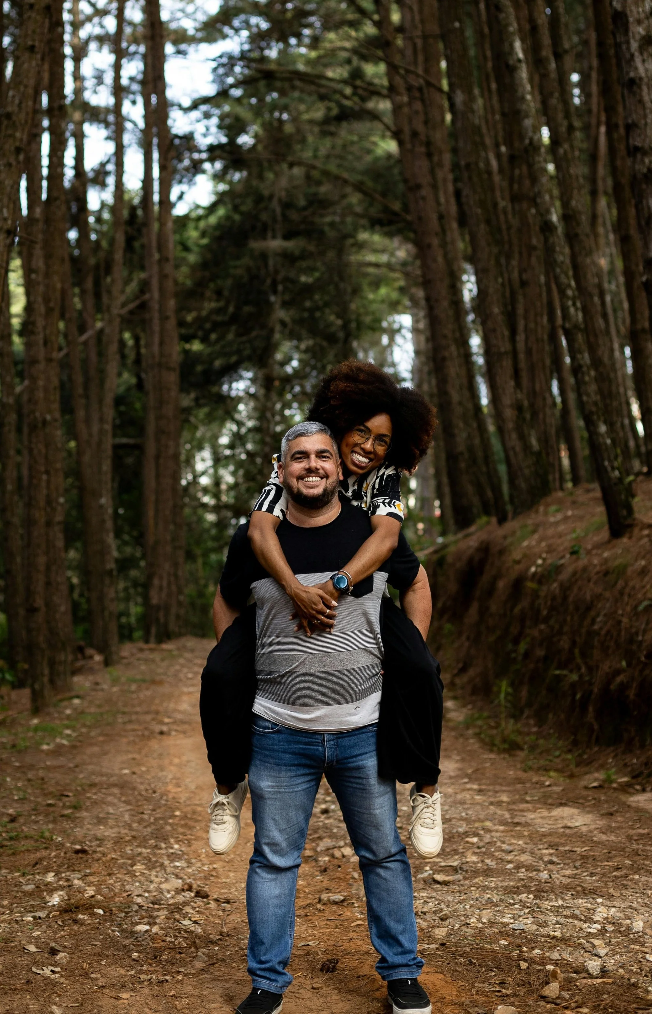 A Latino man stands on a path in the woods holding a black woman piggyback. They are smiling Therapy for interracial couples in Los Angeles