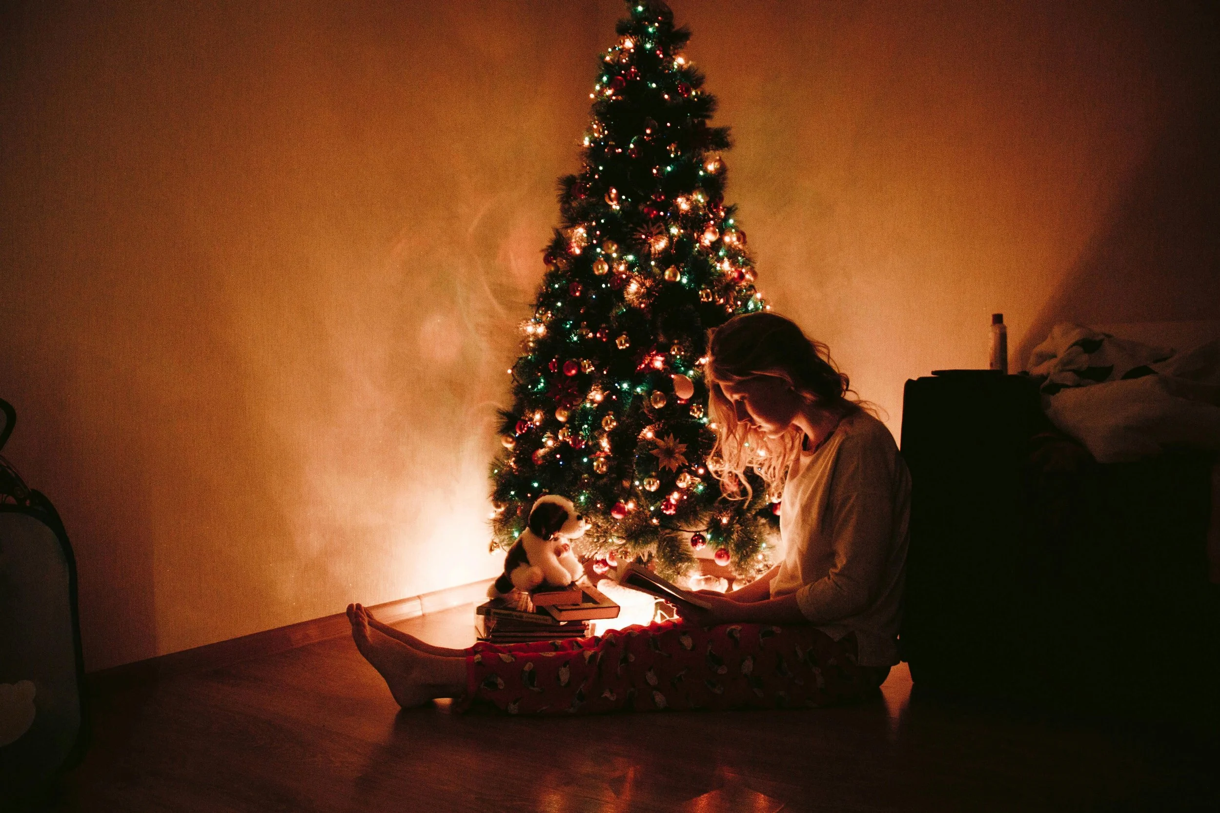 A woman sitting alone by a glowing Christmas tree, reading in soft holiday light, capturing the quiet emotion of being single during the holidays.