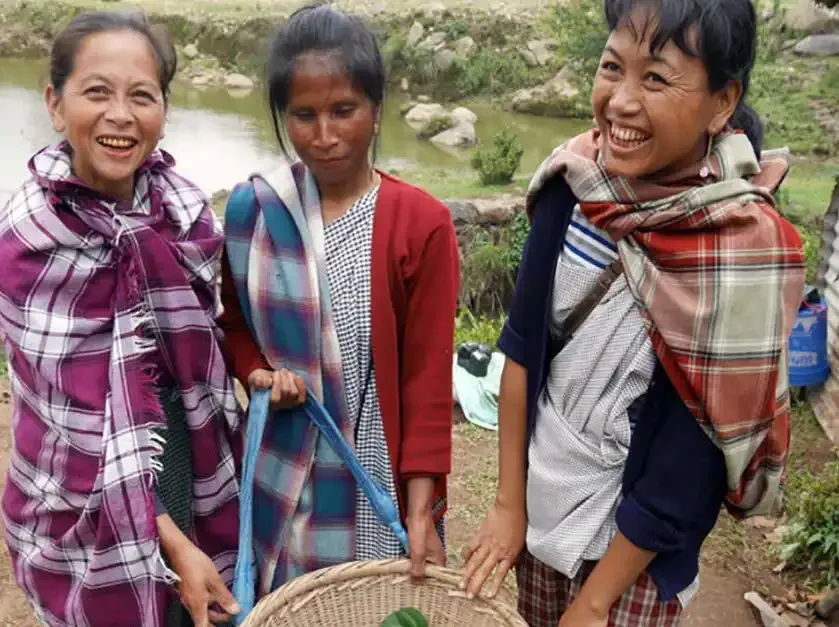 Three smiling women outdoors near a river, carrying a basket with green leaves in the Khasi Hills.