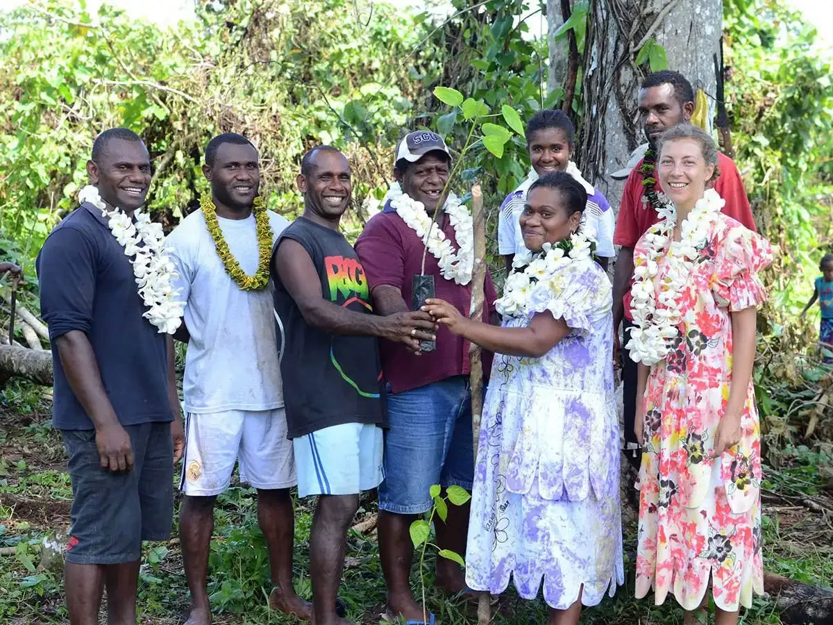 Group of eight people standing outdoors in Loru Forest on planting day, wearing floral garlands, with some holding a small plant and a piece of tree bark.
