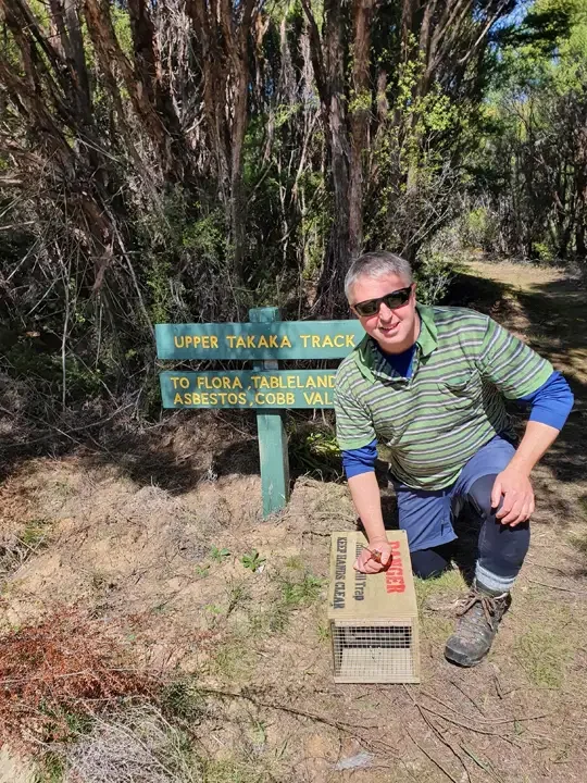 A person outdoors in a forest, crouching next to a metal grating and a trail sign that reads "Upper Takaka Track to Flora, Tableland, Asbestos, Cobb Valley." They are wearing sunglasses, a green striped jacket, and hiking boots.