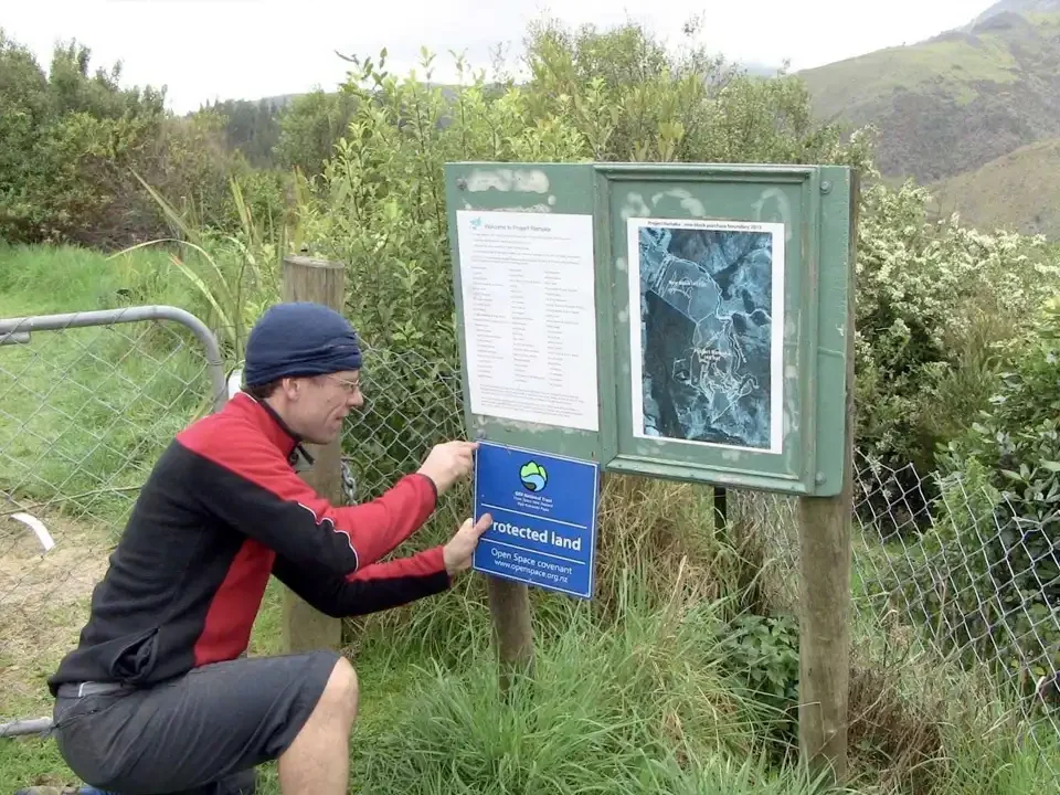 Man attaching a sign to a fence at in Rameka, with informational signs and a map on post, surrounded by greenery and mountains in the background.