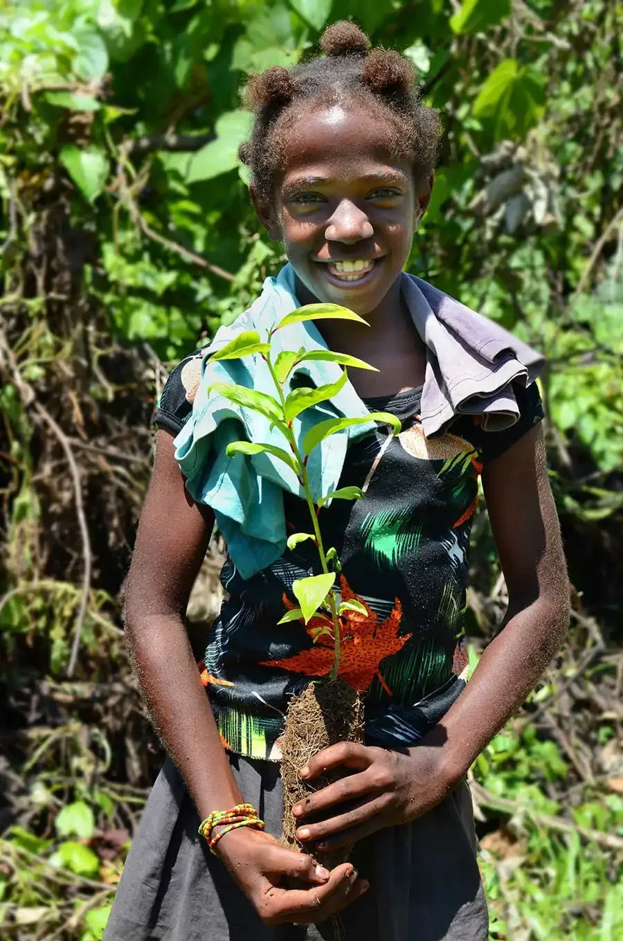 A smiling young girl with braided hair holding a small tree sapling in Loru Forest