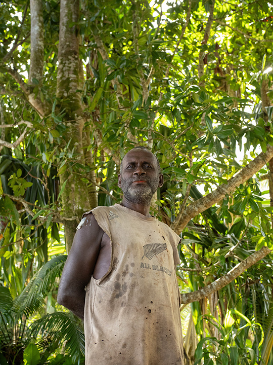 An older man with a beard standing outdoors in a lush, green forested area, wearing a worn sleeveless shirt.