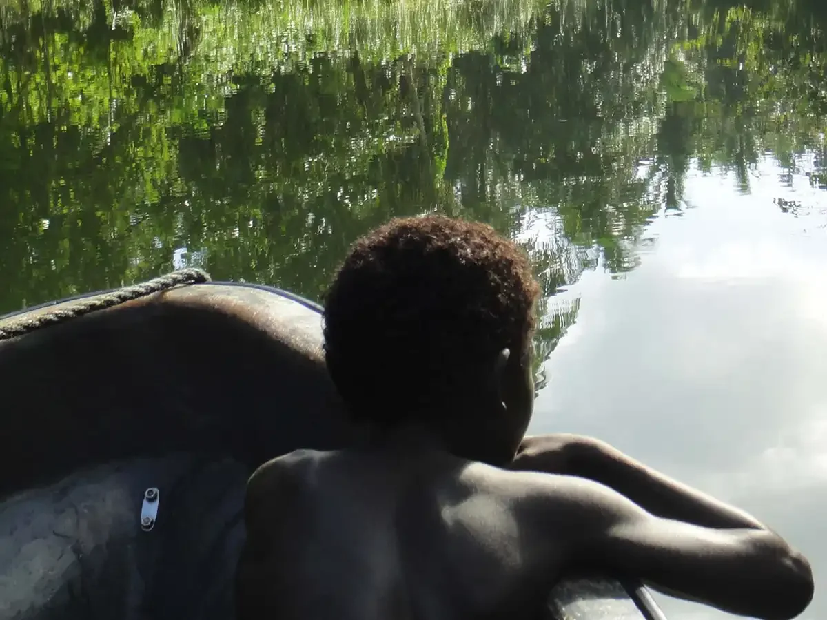 A young man relaxing on a small boat on the Choiseul River in Babatana surrounded by lush green trees and reflected in the water.