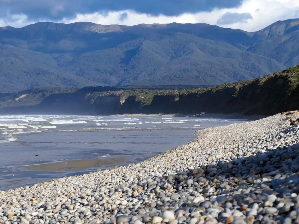 Rarakau beach - a rocky shoreline with small stones, ocean waves, high cliffs, and mountainous terrain in the background under a partly cloudy sky.