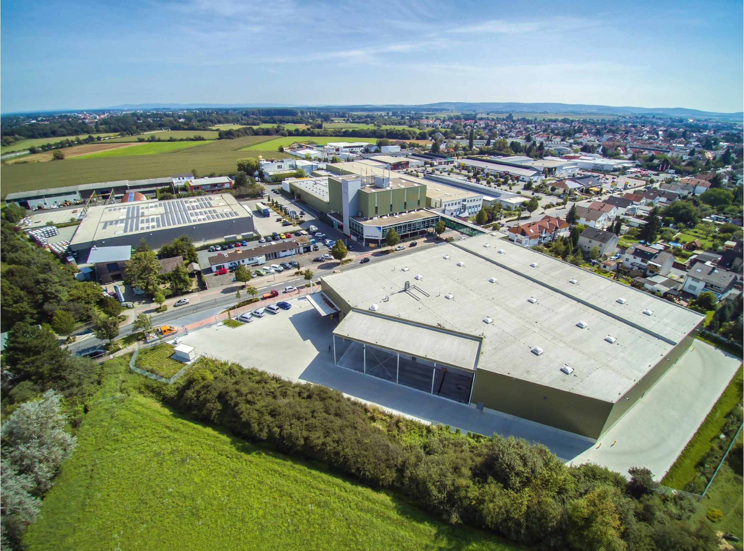 Aerial view of a commercial/industrial area with large buildings, parking lots, surrounded by residential houses and green fields in the background.